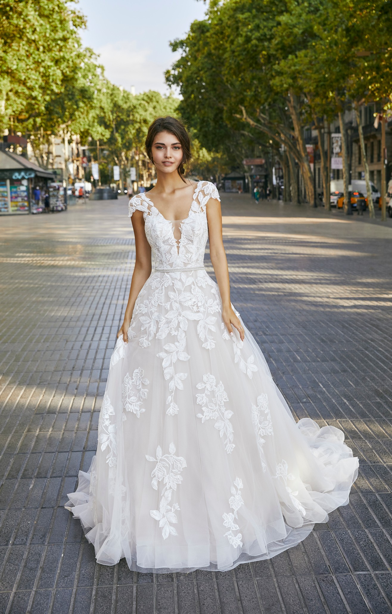 Brunette model stood in a quiet market street in Ronald Joyce 69705, an ivory A-line wedding dress with a plunging illusion v-neckline, bold lace appliqué detailing and a delicately-beaded sparkle belt.