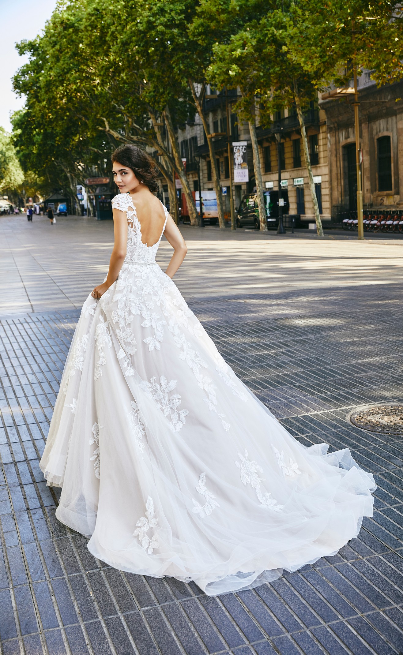 Close up of a brunette model stood in an empty tree-lined street in Ronald Joyce 69705, an ivory A-line wedding dress with a plunging open back, v-neckline, lace applique cap sleeves, bold floral lace appliqués and a delicate sparkly belt.