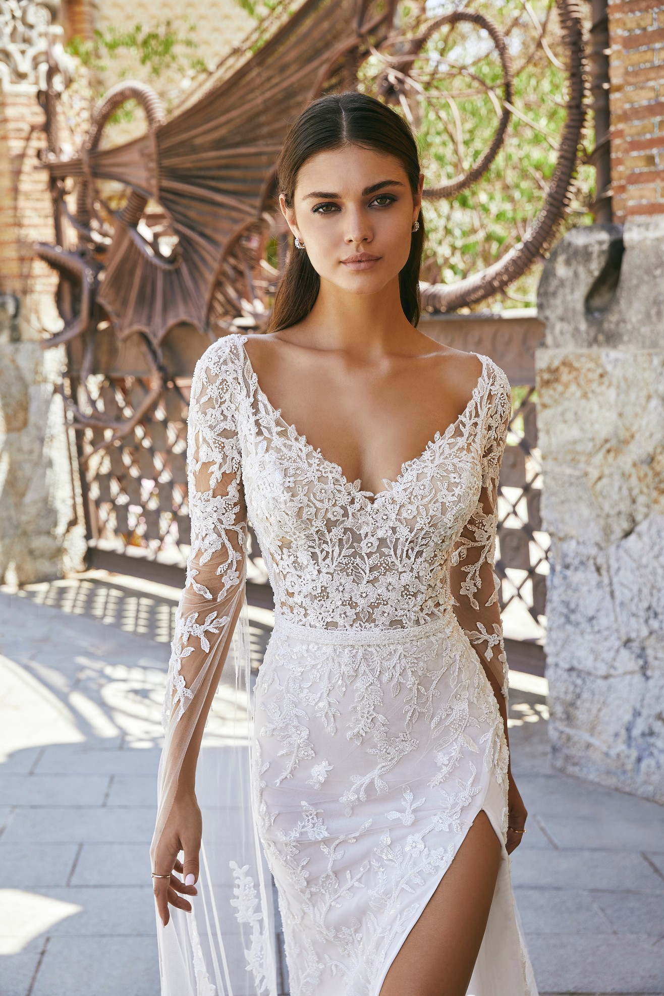 Close up of a model stood in a sunny Italian street in Ronald Joyce style 69706, a sexy lace wedding dress with sheer off-the-shoulder long cape sleeves, an illusion bodice and leg split