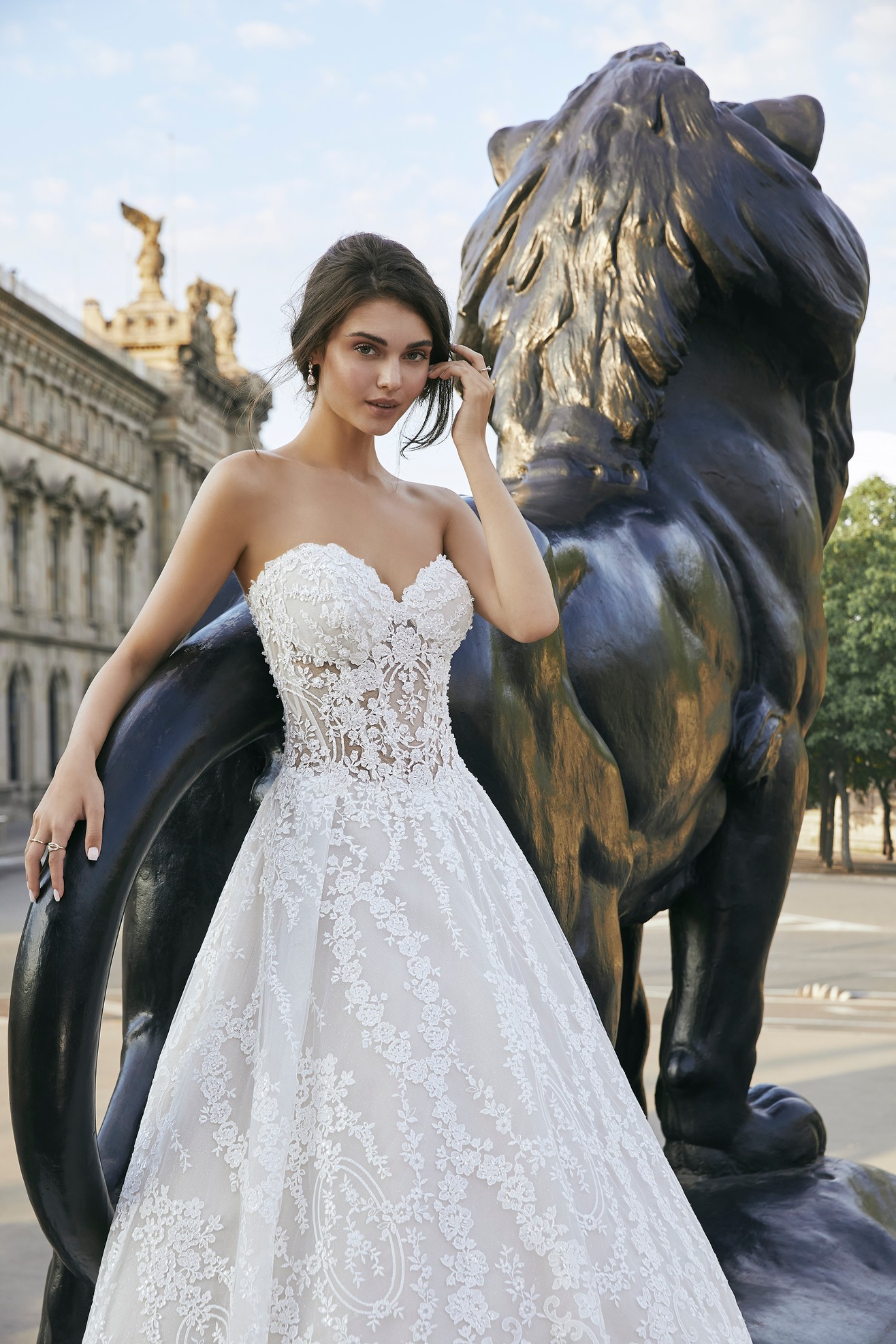 Model stood by a lion statue in Ronald Joyce style 69709, a strapless lace A-line wedding dress with a sweetheart neckline and illusion bodice. 