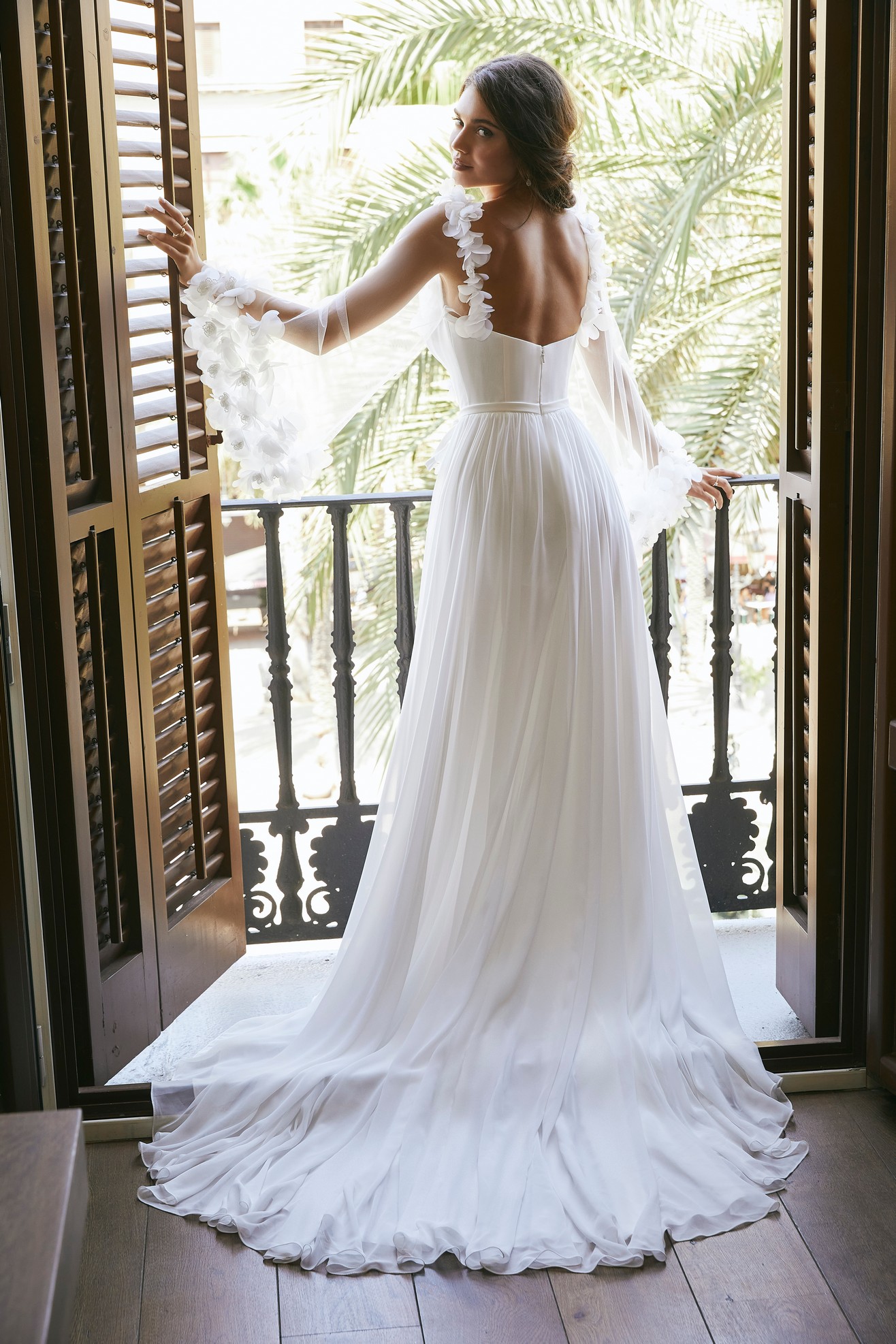 Back of a brunette model stood on a balcony by exotic trees in Ronald Joyce 69710, a chiffon wedding dress with 3D flowers, fluted sleeves, an open back and thigh leg split