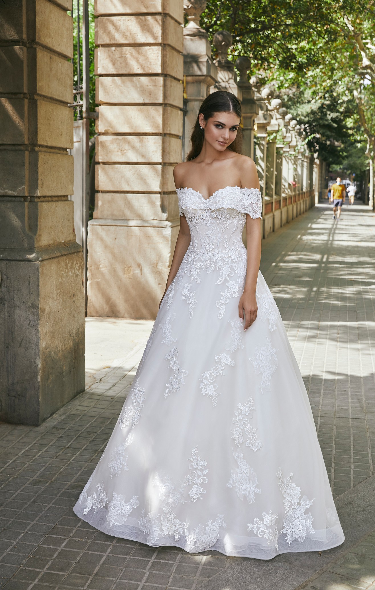 Model stood outside in Ronald Joyce style 69711, an off-the-shoulder lace appliqué A-line wedding dress in ivory.