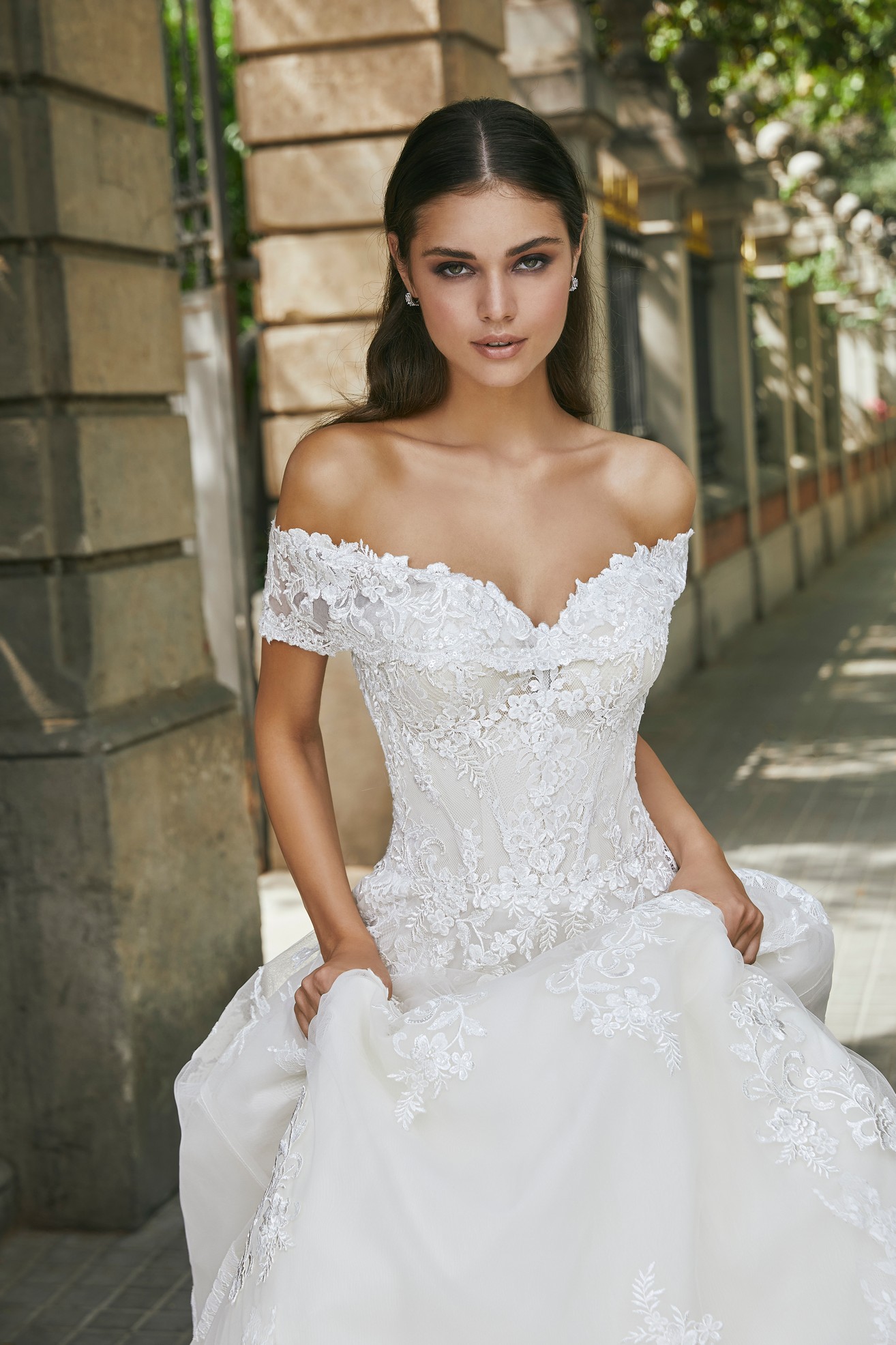 Model stood outside holding the skirt of Ronald Joyce style 69711, an off-the-shoulder lace appliqué A-line wedding dress in ivory.