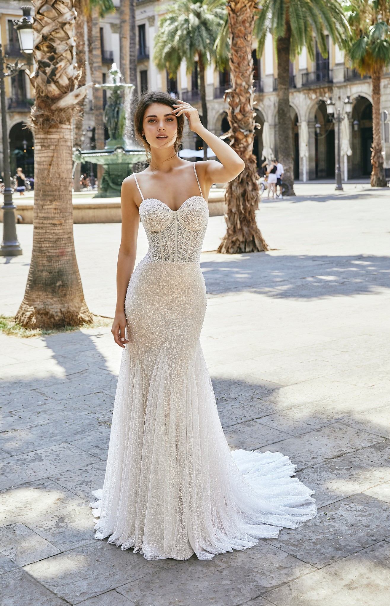 A brunette model stood in a sunny street by exotic trees in Ronald Joyce 69712, an ivory and champagne exquisitely pearl-beaded fit and flare wedding dress with delicate straps and glitter tulle