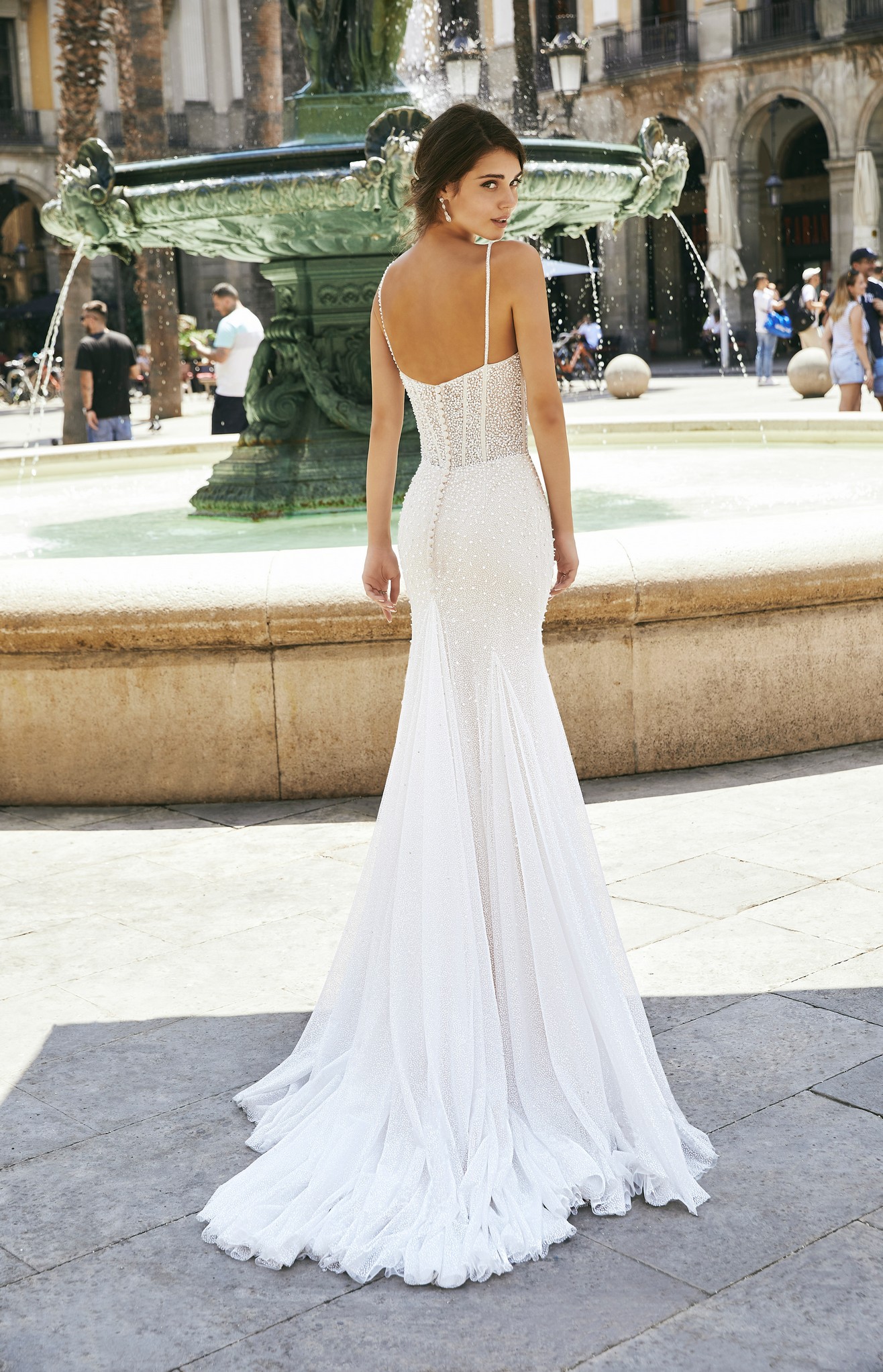 Back of model stood by a fountain in Ronald Joyce style 69712, a pearl-beaded fit and flare wedding dress in glitter tulle with delicate straps and a sweetheart neckline