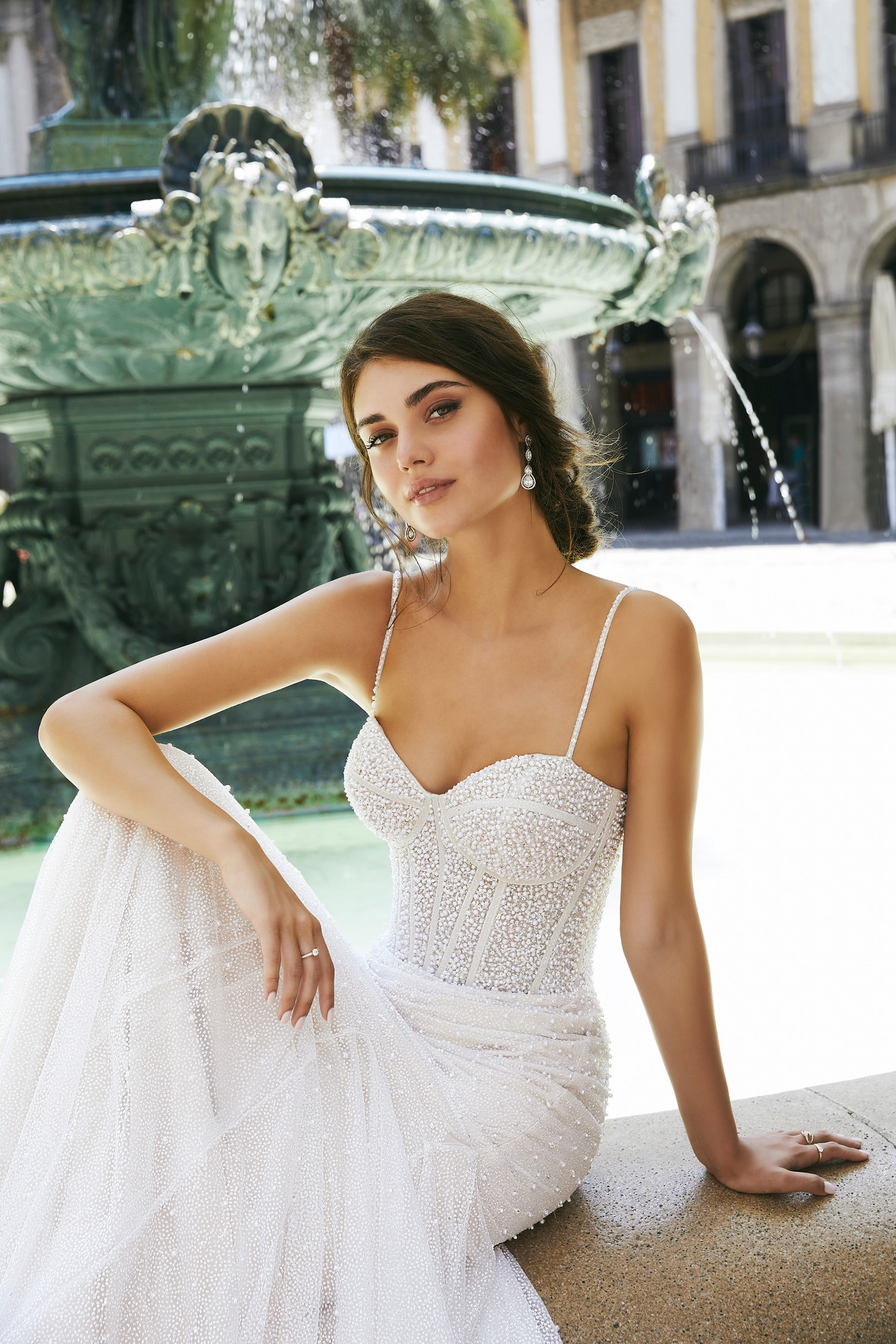 Close up of a brunette model sat by a sunny street water fountain in Ronald Joyce 69712, a sparkly pearl and crystal-beaded fit and flare wedding dress in glitter tulle with delicate straps and a sweetheart neckline
