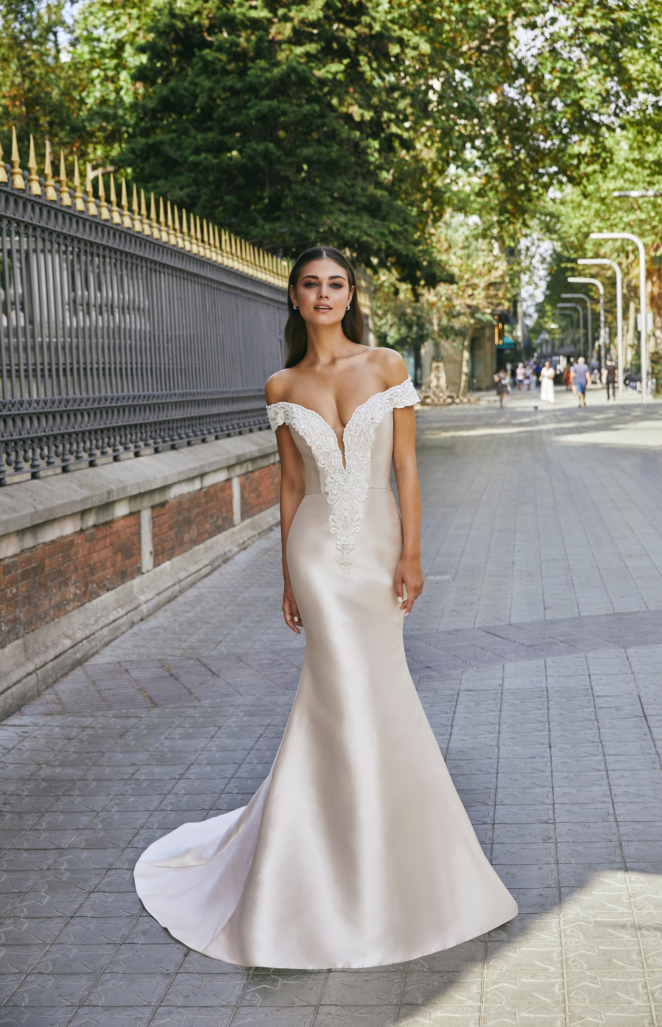 A brunette model stood by black and gold railing in Ronald Joyce 69713, a light gold fit and flare Mikado wedding dress with a plunging off-the-shoulder neckline