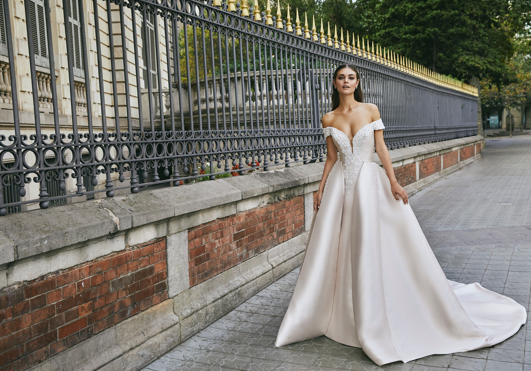 A brunette model stood by black railing in Ronald Joyce 69713, a light gold off-the-shoulder fit and flare wedding dress with a plunging neckline and A-line overskirt made from Mikado bridal fabric