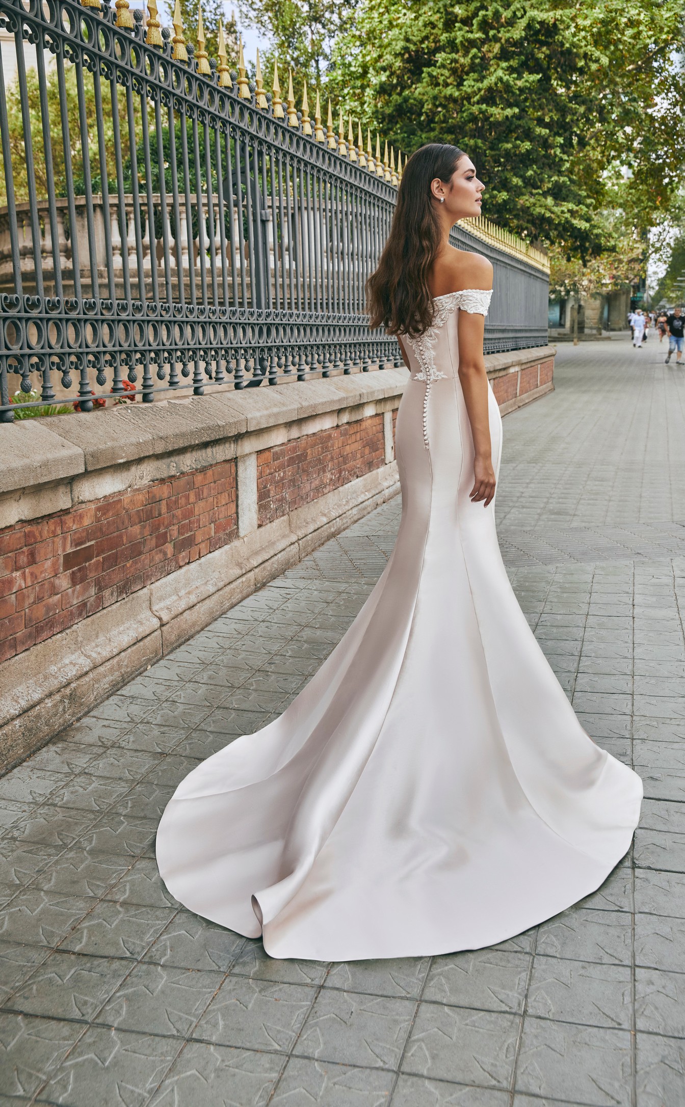 The back of a brunette model stood by posh black railing in Ronald Joyce 69713, a light gold fit and flare Mikado wedding dress with off-the-shoulder ivory lace applique sleeves