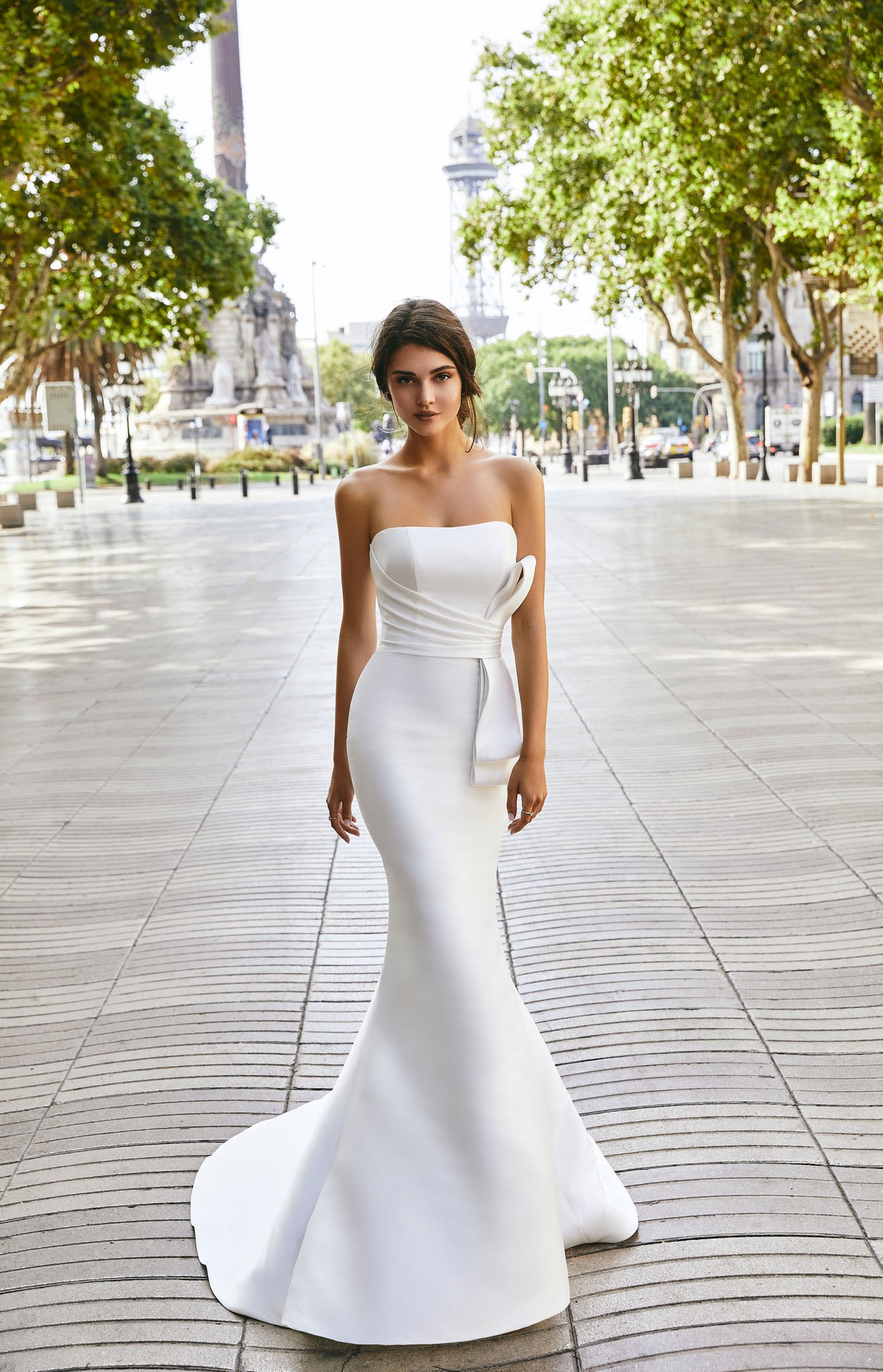 Model stood in a street wearing Ronald Joyce 69714, a strapless classic fit and flare wedding dress with a statement bow and bridal buttons cascading down the dress to the hemline.