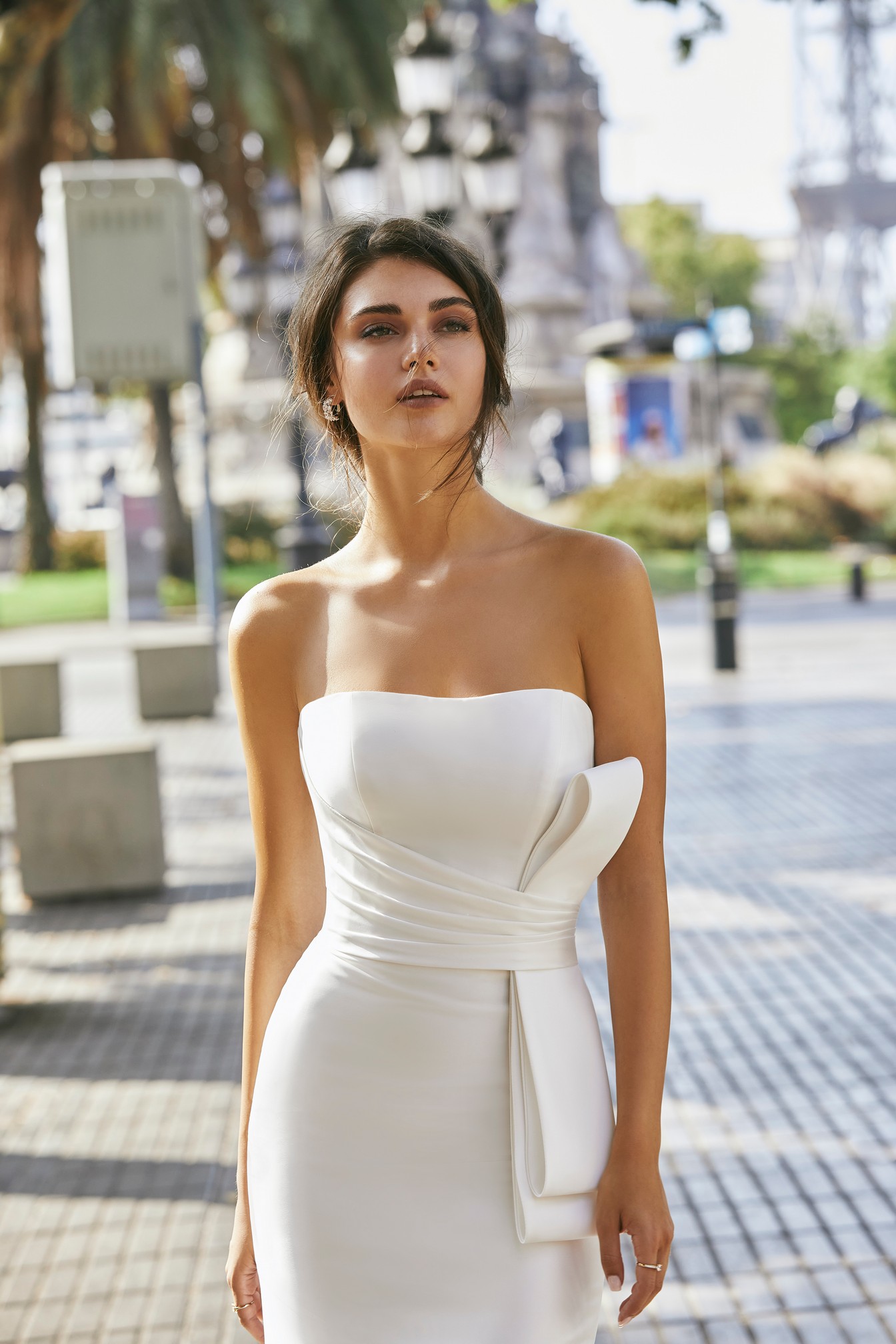 Close up of model stood in a street wearing Ronald Joyce 69714, a plain strapless fit and flare wedding dress with a statement bow.