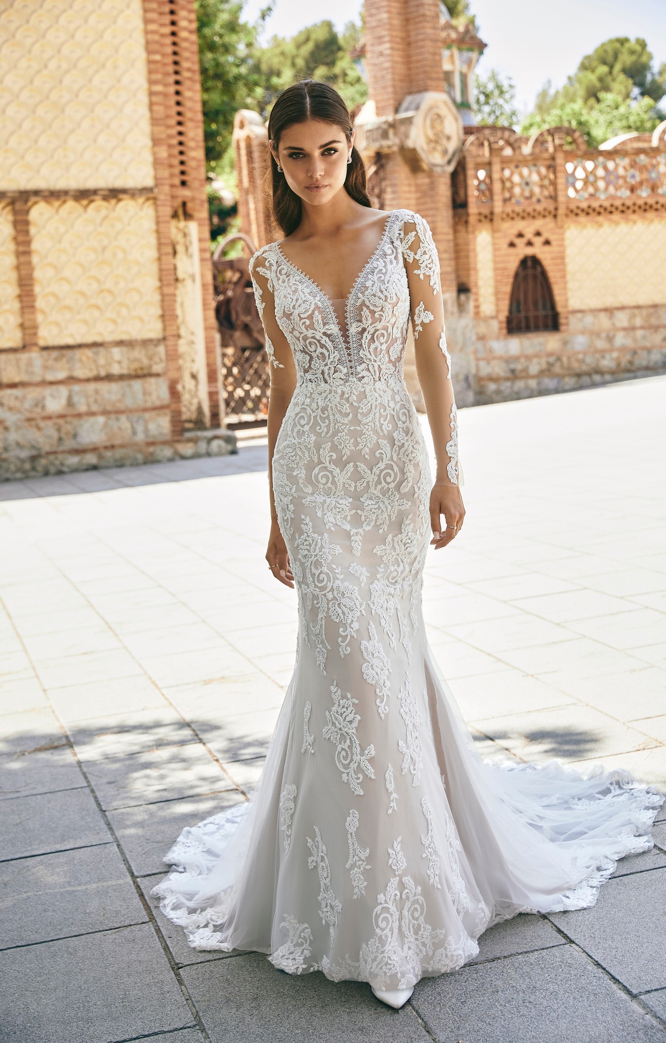 A close up of a brunette model stood in a sunny street in Ronald Joyce 69716, a tulle and lace applique bridal fabric wedding dress with long illusion lace sleeves and a plunging neckline