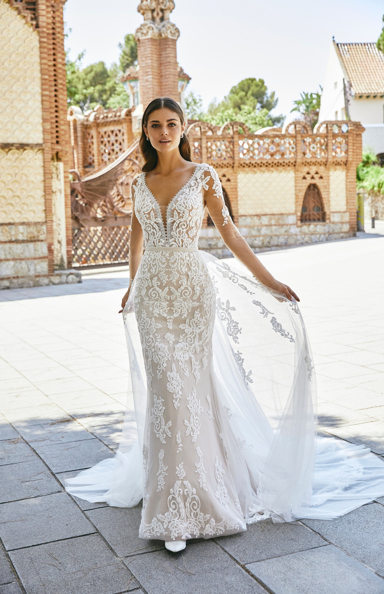 Front profile of model wearing Ronald Joyce wedding dress style 69716, a tulle and lace applique fit and flare dress with illusion full sleeves and a plunging illusion neckline and v-shaped back complete with button and bustle detailing. 