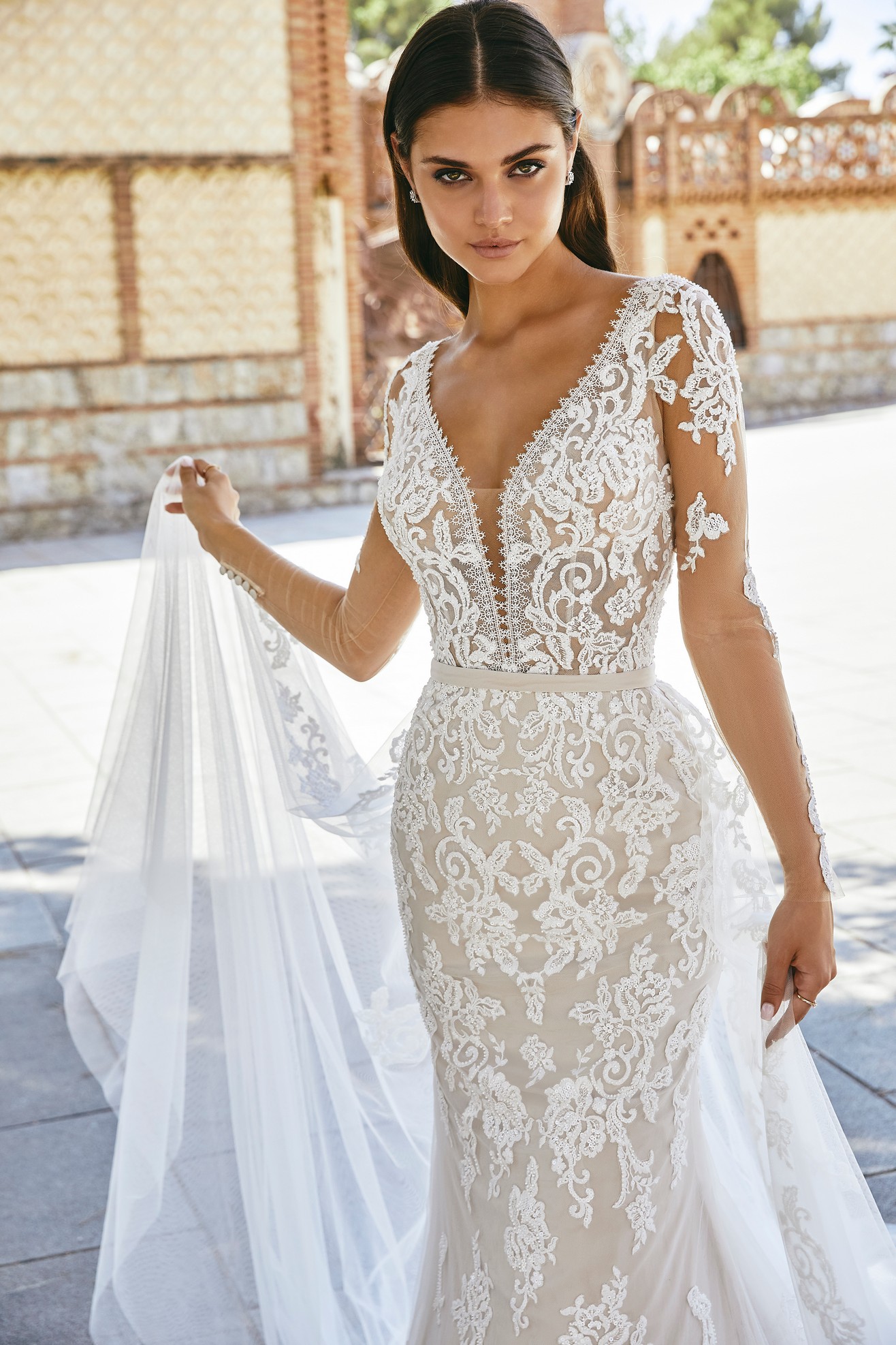 A close up of a brunette model stood in a sunny street in Ronald Joyce 69716, a lace applique wedding dress with long illusion lace sleeves and a plunging illusion neckline