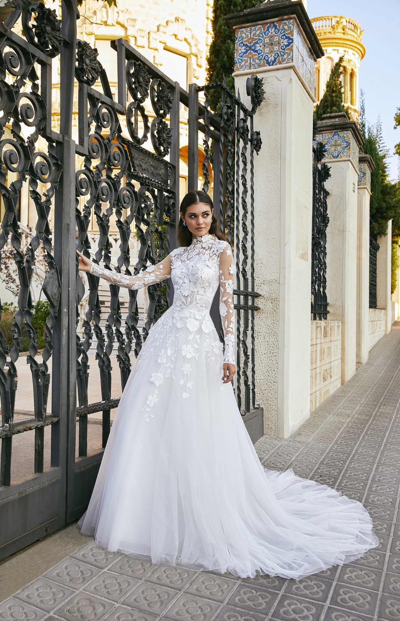 Model stood by black railing in Ronald Joyce style 69717, a high neck A-line wedding dress with lace neckline and sleeves, 3D flowers and a tulle skirt and train