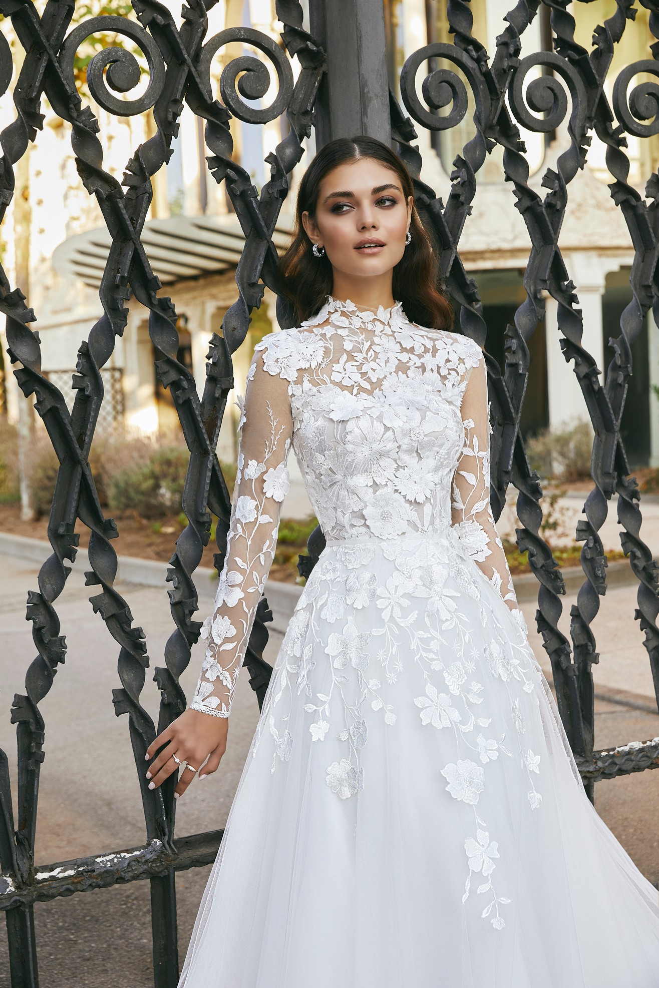 Brunette model stood by black railing in Ronald Joyce 69717, an exquisite lace applique A-line wedding dress with an illusion high neckline, long sheer applique sleeves, 3D flowers and an organza and tulle skirt.