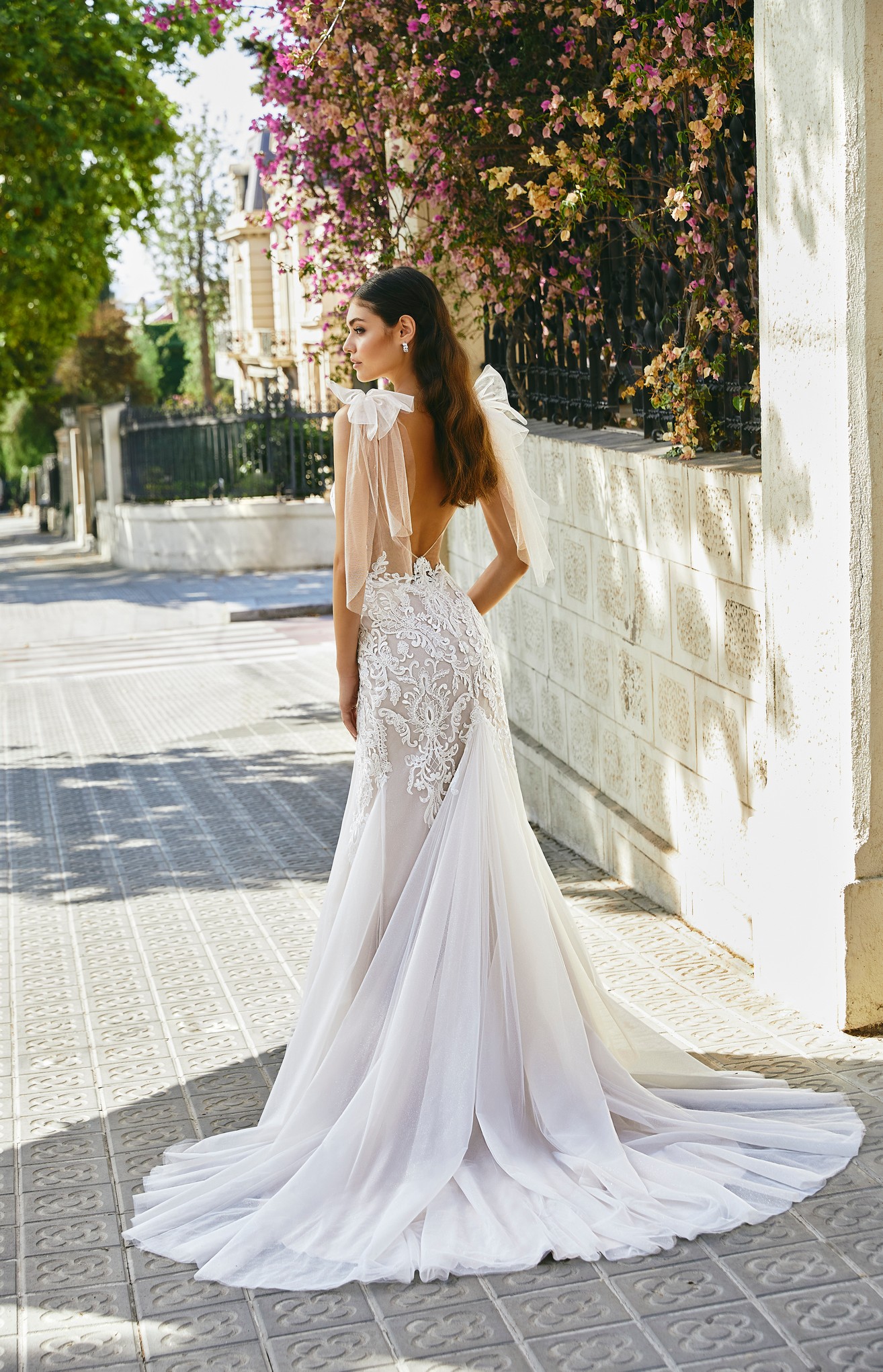 Back of model stood outside in Ronald Joyce style 69719, a glitter tulle and lace fit and flare wedding dress with an open back, statement tulle shoulder bows and a bustle. 
