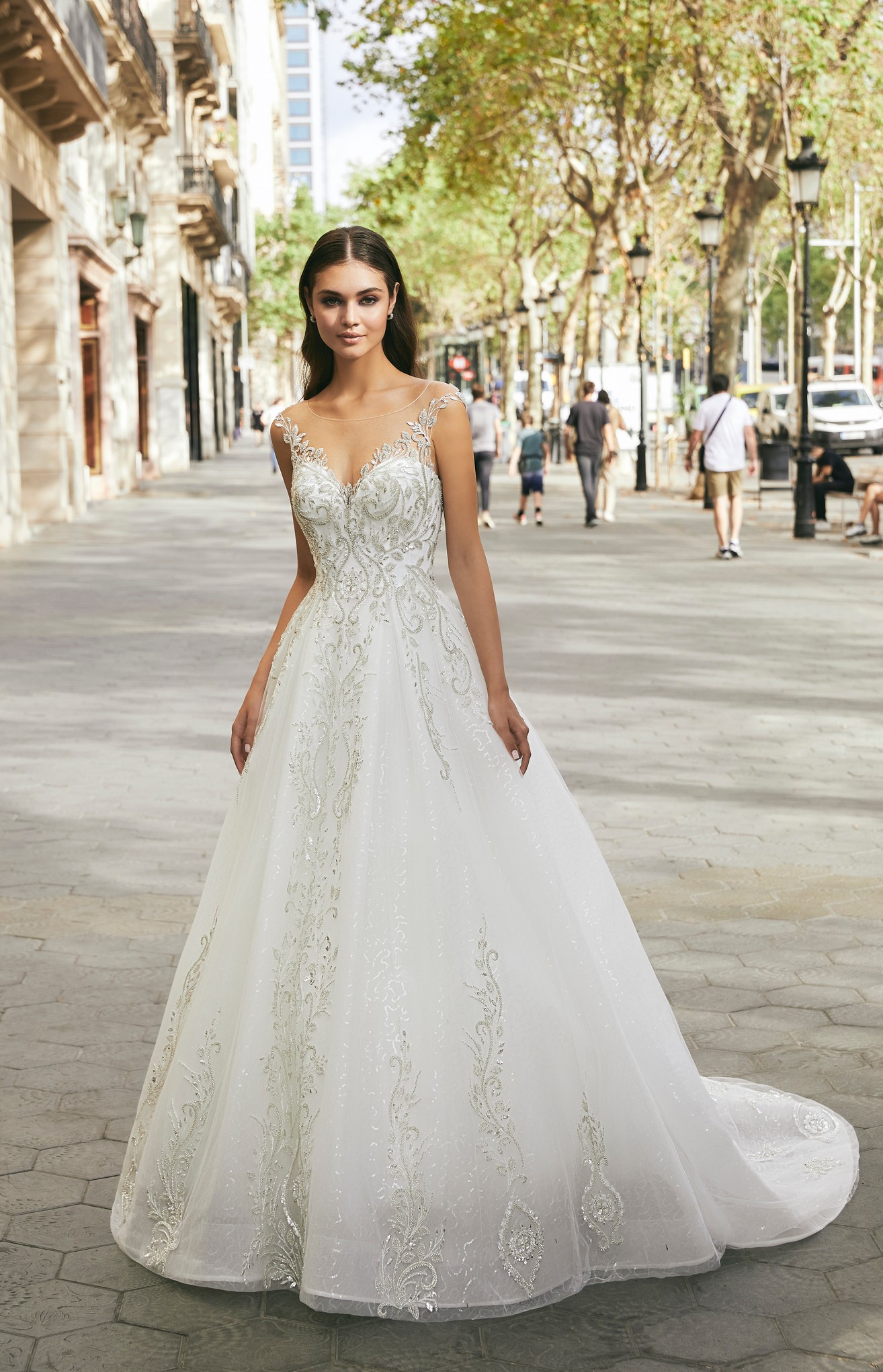 Brunette model stood in a sunny black lamp post and tree-lined street in Ronald Joyce 69720, an ivory and silver sparkly A-line wedding dress with a sweetheart bodice, glitter tulle skirt and sparkly illusion off-the-shoulder sleeve.