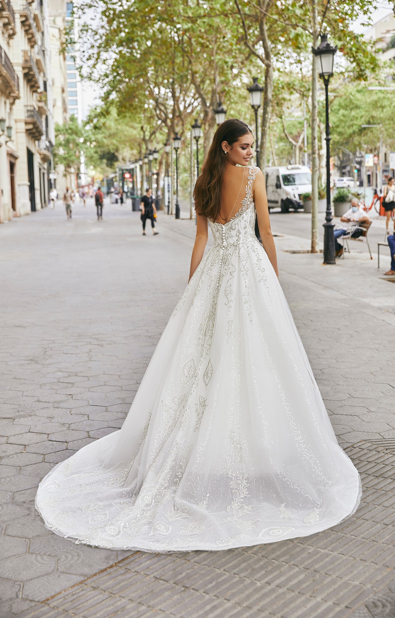 Back of a brunette model stood in a sunny black lamp post and tree-lined street in Ronald Joyce 69720, an ivory and silver sparkly A-line wedding dress with an open back, glitter tulle skirt and sparkly illusion off-the-shoulder sleeve. 