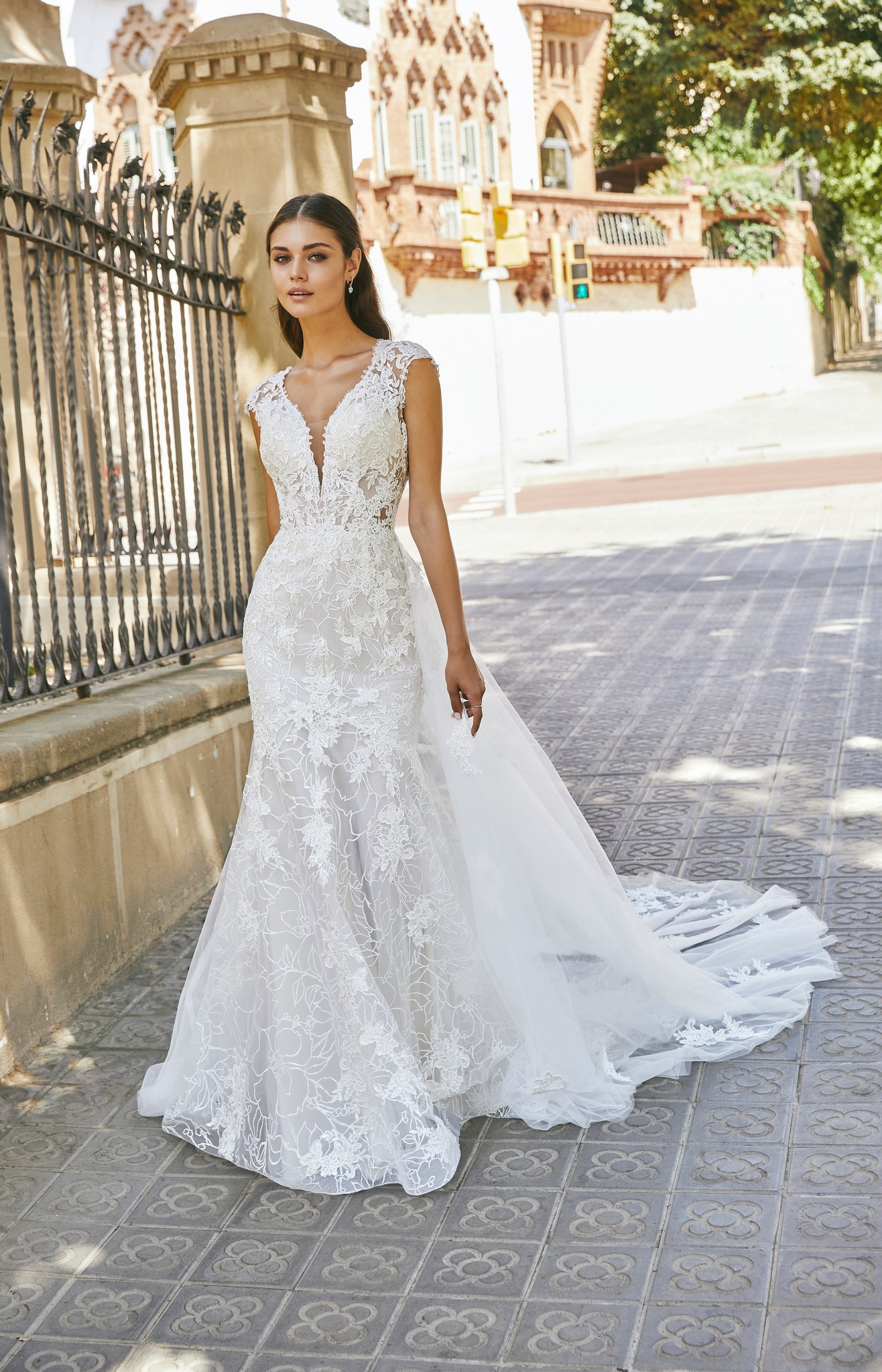 Brunette model stood in a sunny street by black railing in Ronald Joyce 69721, a floral lace and tulle wedding dress style with cap sleeves, a plunging v-neckline and v-shaped open back.
