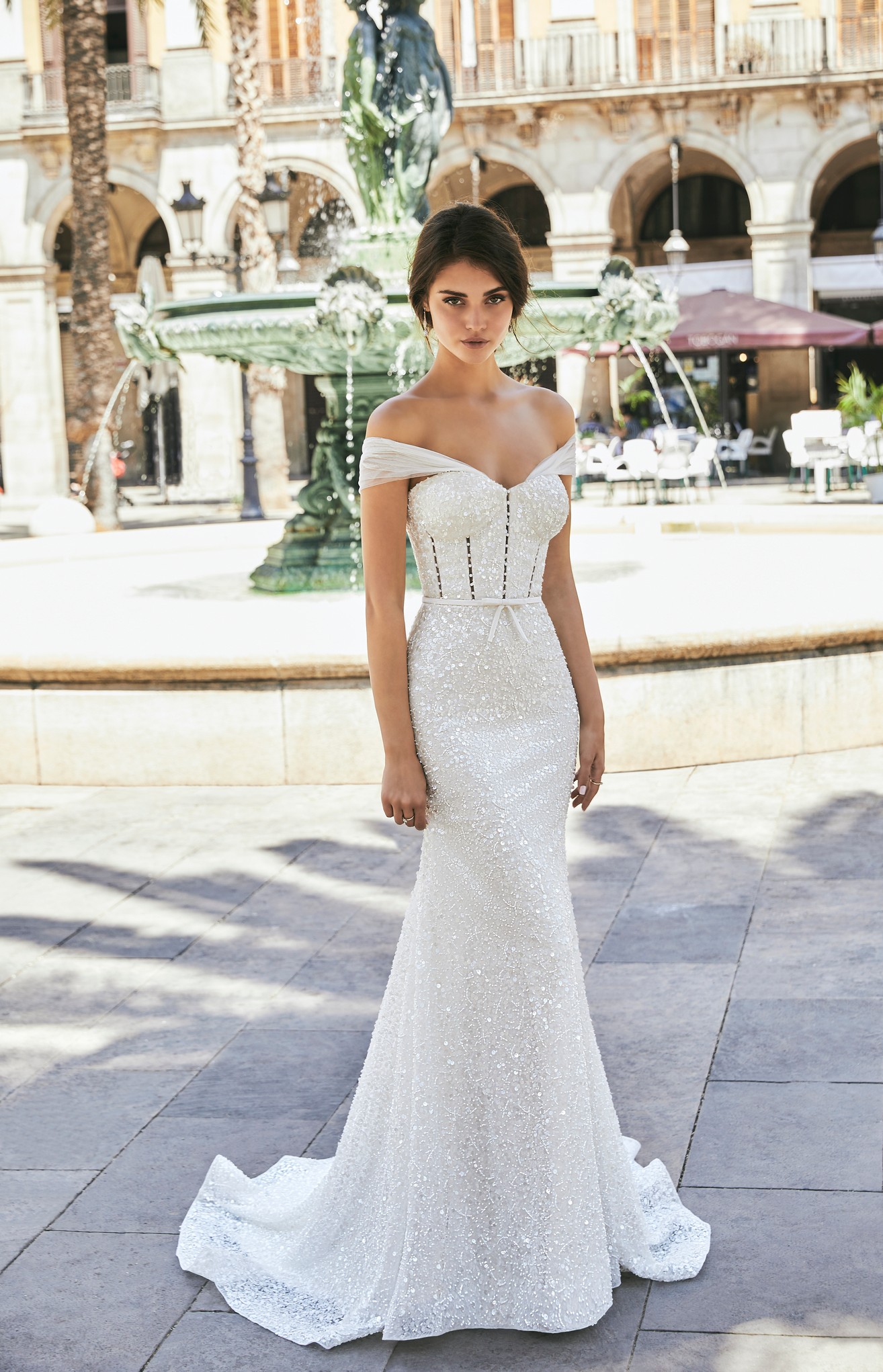 Brunette model stood by a sunny green street fountain in Ronald Joyce 69722, a sparkly fit and flare wedding dress with a pearl-beaded and sequin bodice, sweetheart neckline, delicate bow waist belt and detachable off-the-shoulder sleeves