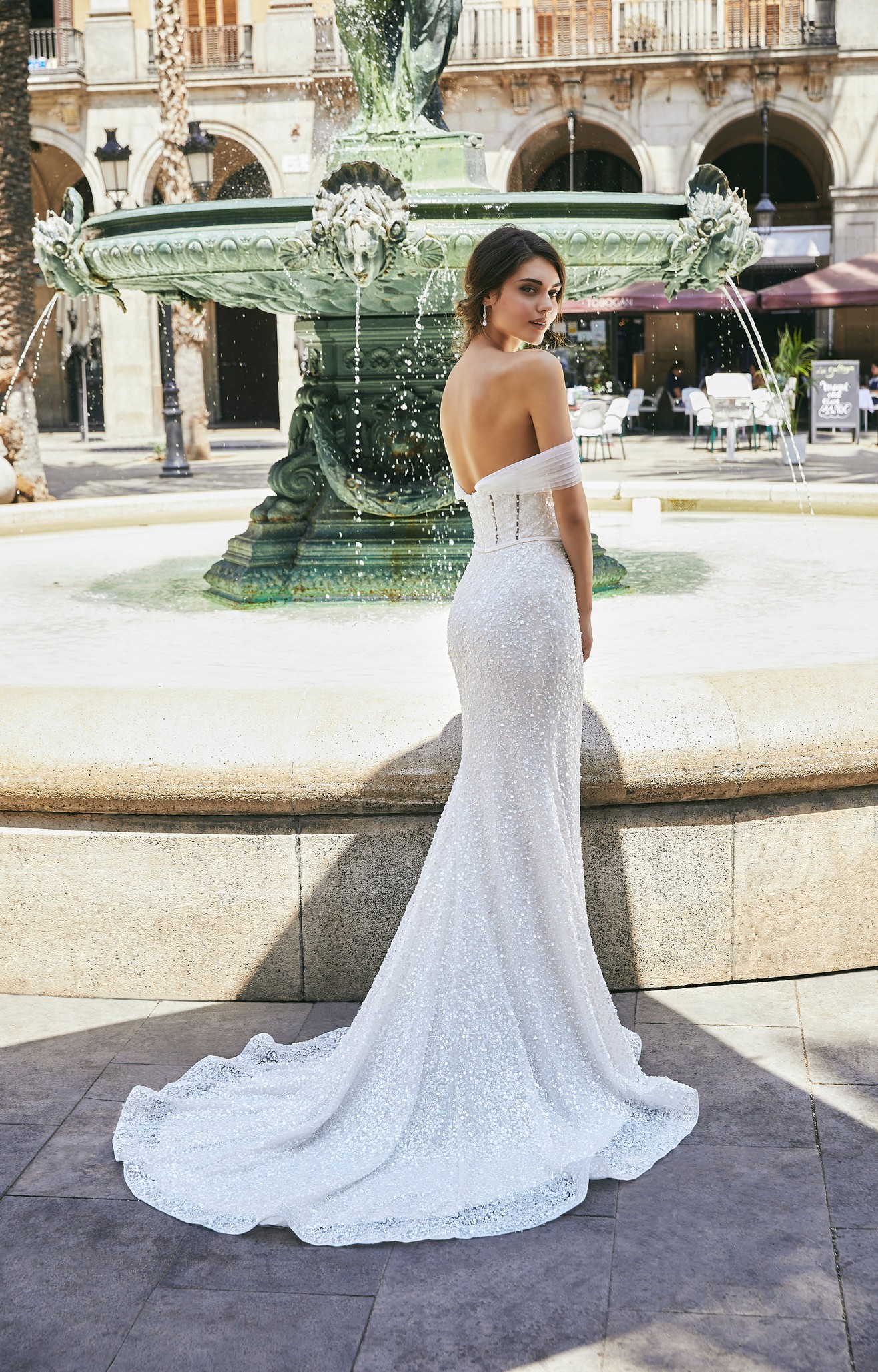 Back of a brunette model stood by a sunny green street fountain in Ronald Joyce 69722, a sparkly fit and flare wedding dress with a pearl-beaded and sequin bodice, open back and detachable off-the-shoulder pleated tulle sleeves