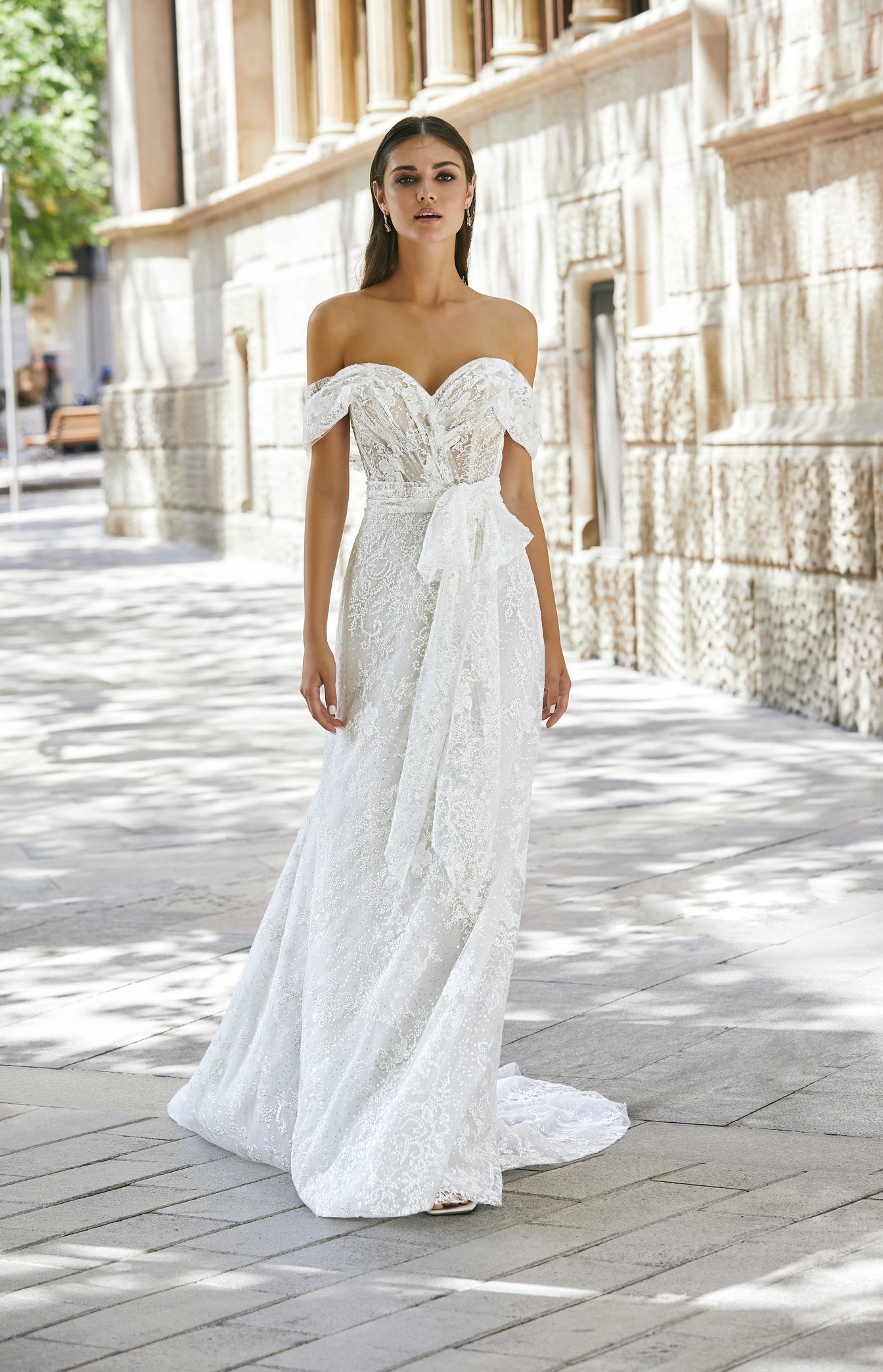 Brunette model stood outside a sunny stone building in Ronald Joyce 69724, a floaty lace  off-the-shoulder wedding dress style with draped cap sleeves, an illusion bodice and striking bow waist detail