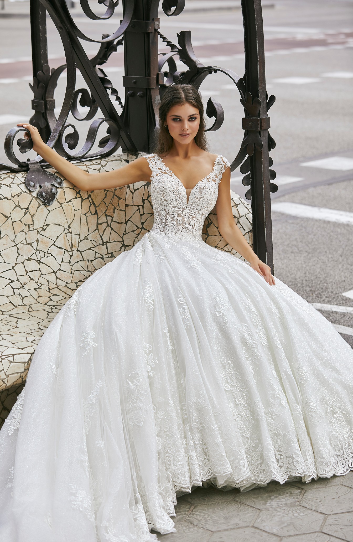 Model sat by black railing in Ronald Joyce style 69726, a beautiful ballgown wedding dress with detachable long illusion sleeves, a glitter and lace tulle skirt and train.