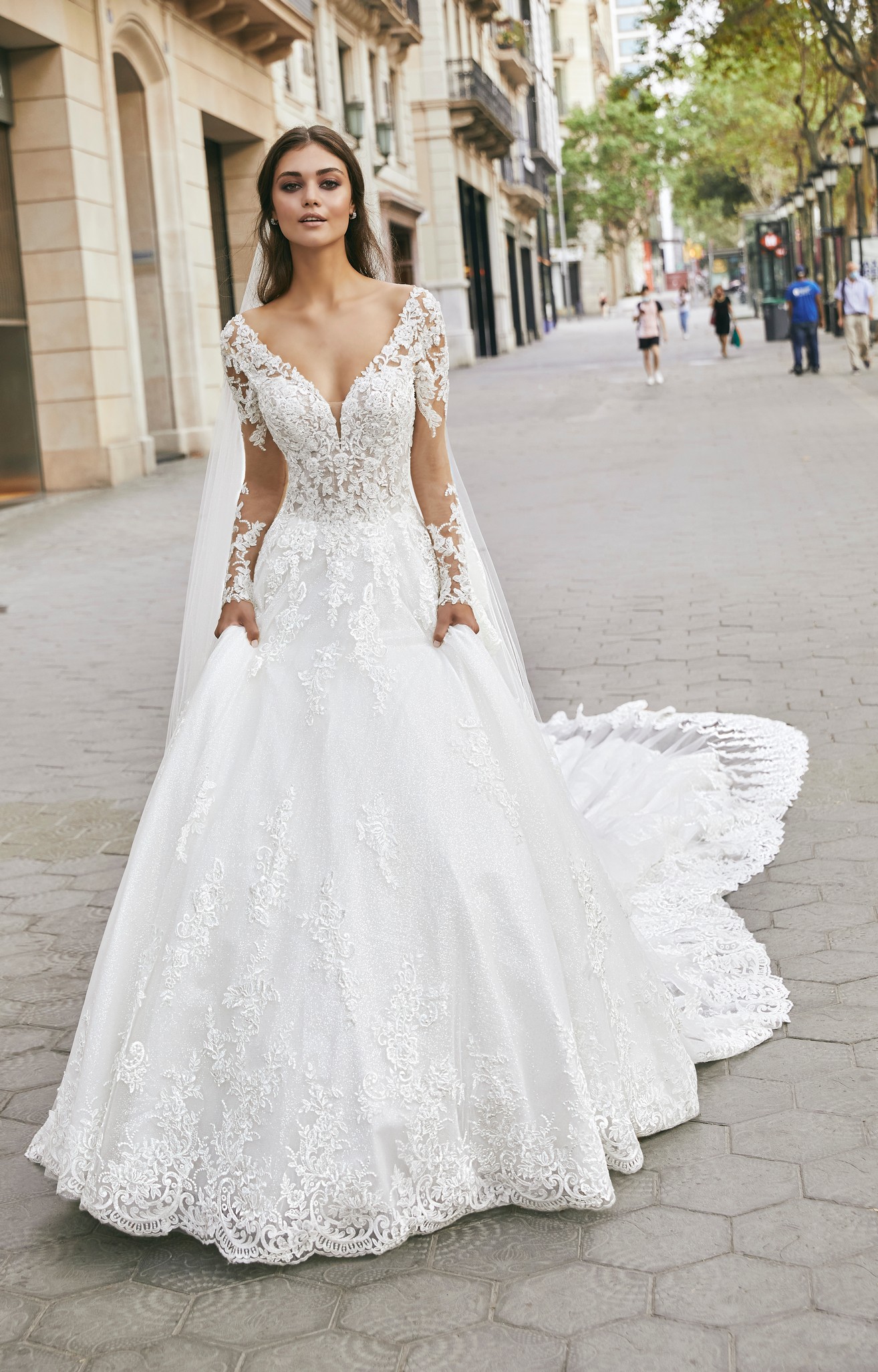 Brown haired lady standing on street in Barcelona wearing long lace sleeved ballgown wedding dress and veil