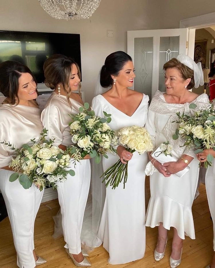 Close up of real bride Emma stood inside with 2 bridesmaids and her mum. The bridemaids wear jumpsuits with white trousers and gold tops. Emma wears her white Ronald Joyce Tai wedding dress. Mum wears an ivory hi low dress.