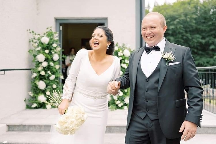 Real bride Emma beaming with happiness, stood outside holding hands with husband Brian in Ronald Joyce Tai 18202, a fitted white wedding dress with long sleeves and a v-neckline.