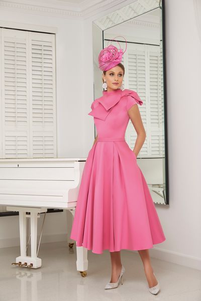 Model stood in a cream room by a white piano in Veni Infantino 991639, modern candy pink Mikado A -line dress with cap sleeves, a statement shoulder bow and hidden pockets