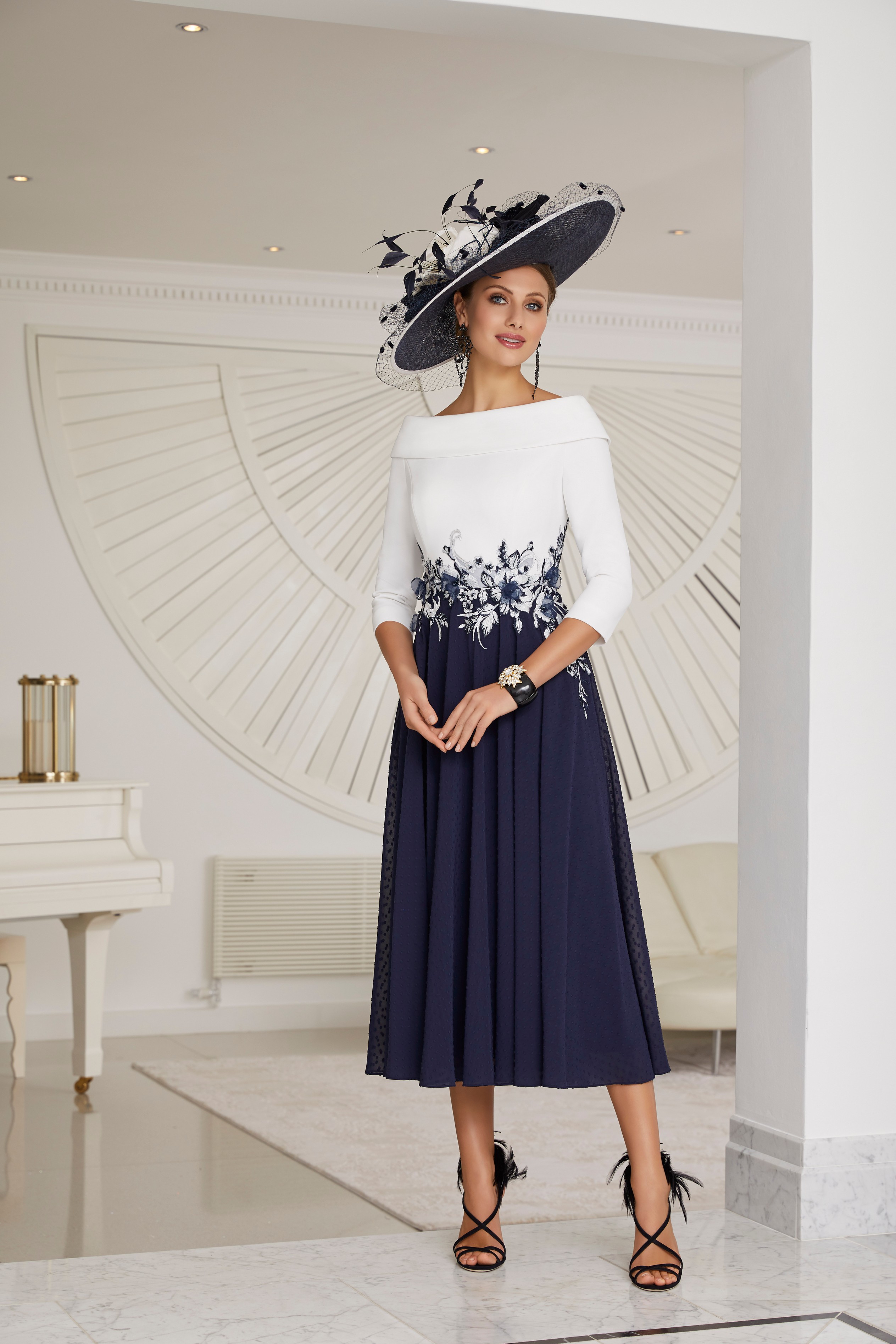 Model stood in an all cream room wearing a navy and ivory hat and Veni Infantino 991654, a navy and ivory calf-length A-line dress with floral lace waist detail, a bateau neckline and ¾ sleeves