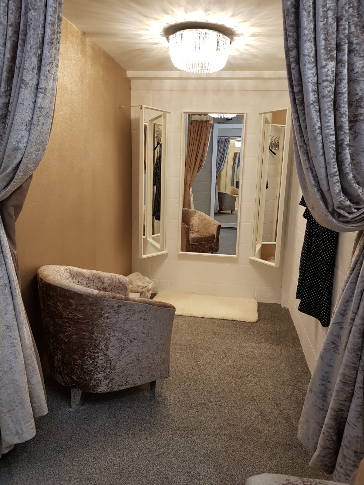 The dressing room at Simply Wed Bridal in Newcastle complete with gold wall, triptych mirror, grey plush chair and matching curtains, grey carpet and glass light