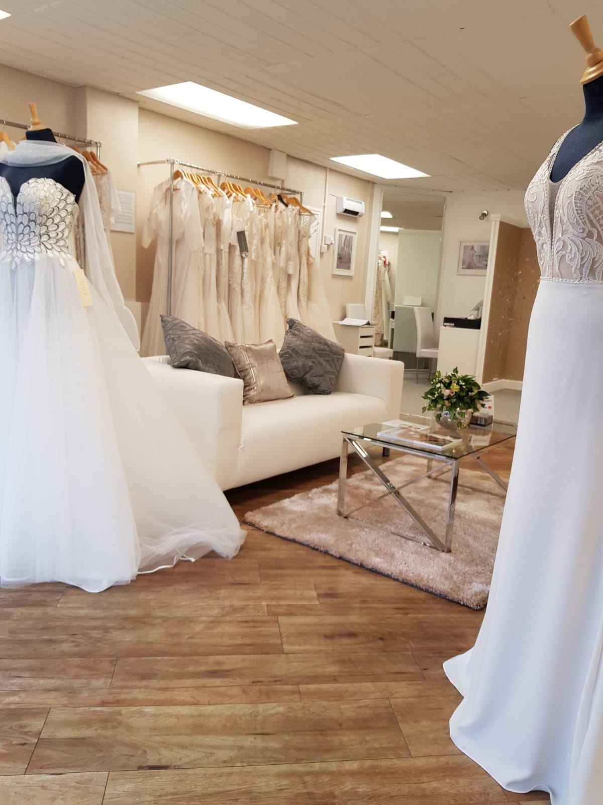 Newcastle's Simply Wed Bridal’s spacious waiting room complete with glass coffee table, pink rug, cream sofa, cushions, mannequins and wedding dresses neatly hung on a rail