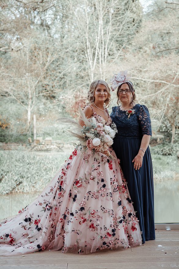Ronald Joyce real bride, Rebecca, and her mum standing by a lake. Rebecca has curled blonde hair and wears Celestina, our colourful ballgown wedding dress with bold floral appliques. Her mum wears a long navy dress with a lace bodice and ¾ sleeves. 