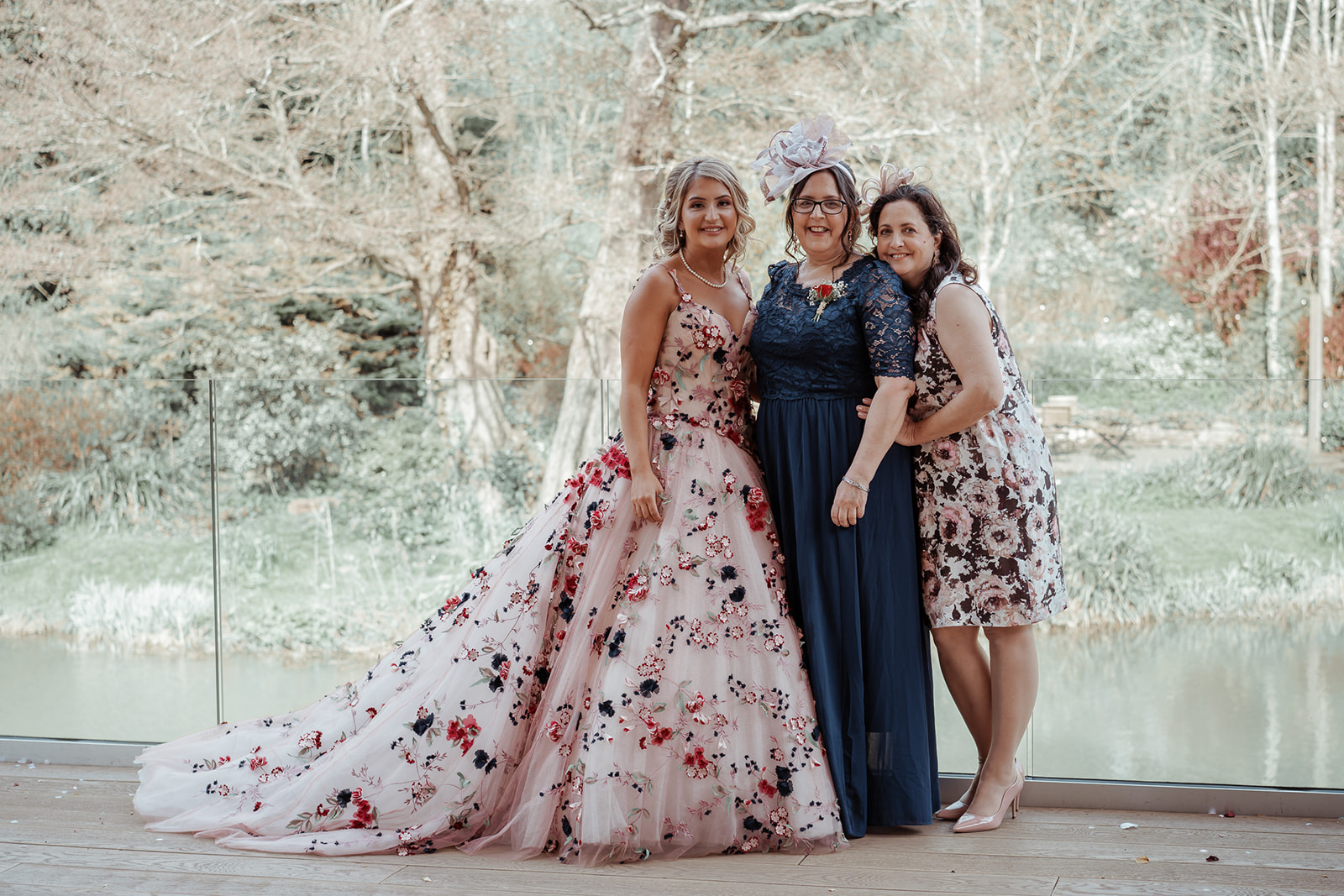 Ronald Joyce real bride, Rebecca, and two female guests standing by a lake. Rebecca has curled blonde hair and wears Celestina, our colourful ballgown wedding dress with claret, navy and green floral appliques.