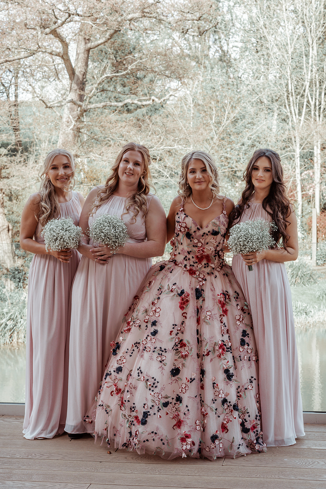 Ronald Joyce real bride, Rebecca, and her three bridesmaids standing by a lake. Rebecca has curled blonde hair and wears Celestina, our colourful ballgown wedding dress with bold floral appliques. Her bridesmaids wear long pink dresses. 