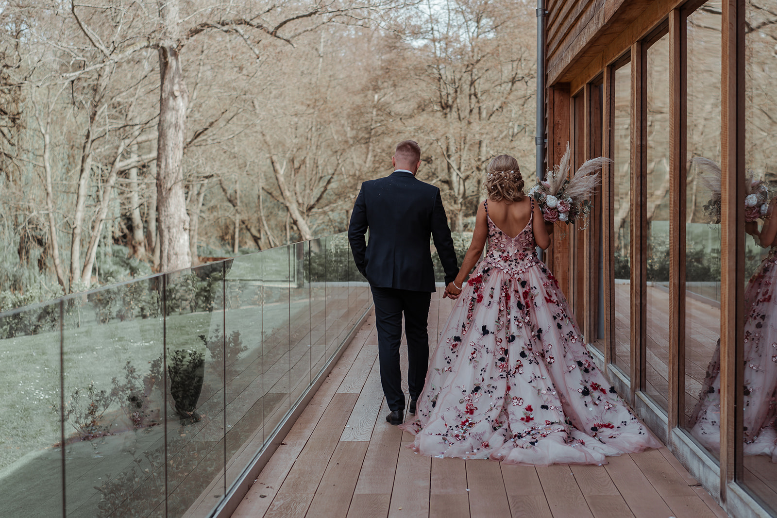 Ronald Joyce real bride, Rebecca, and her husband, Michael, holding hands and walking outside on their wedding day. Rebecca has curled blonde hair and wears our Celestina ballgown wedding dress, a colourful ballgown with coloured floral appliques.