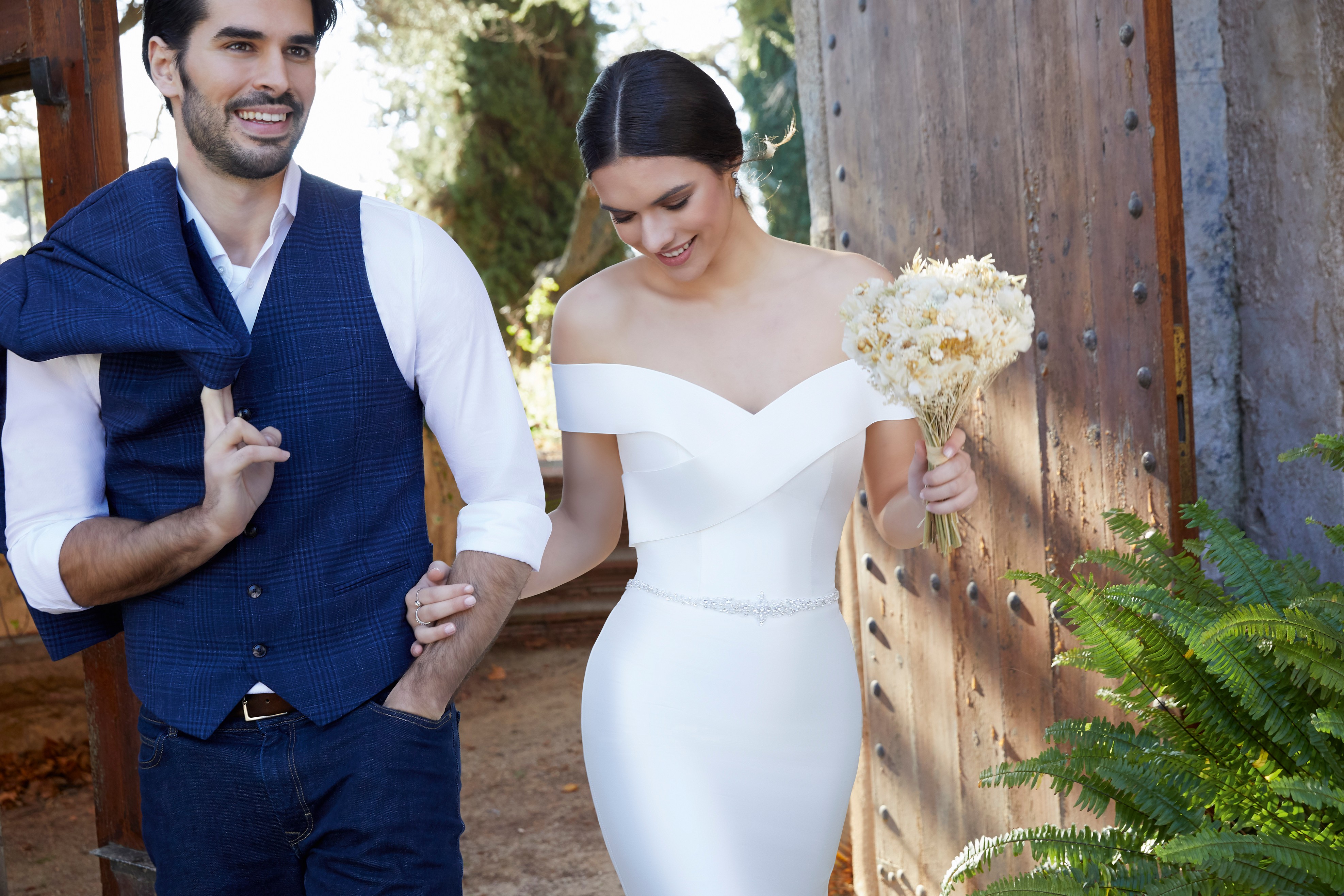Model and groom stood arm in arm by a gate. Model wears a bardot wedding dress (Ronald Joyce 18452), an off the shoulder plain Mikado fit and flare dress in ivory with a sparkle belt.