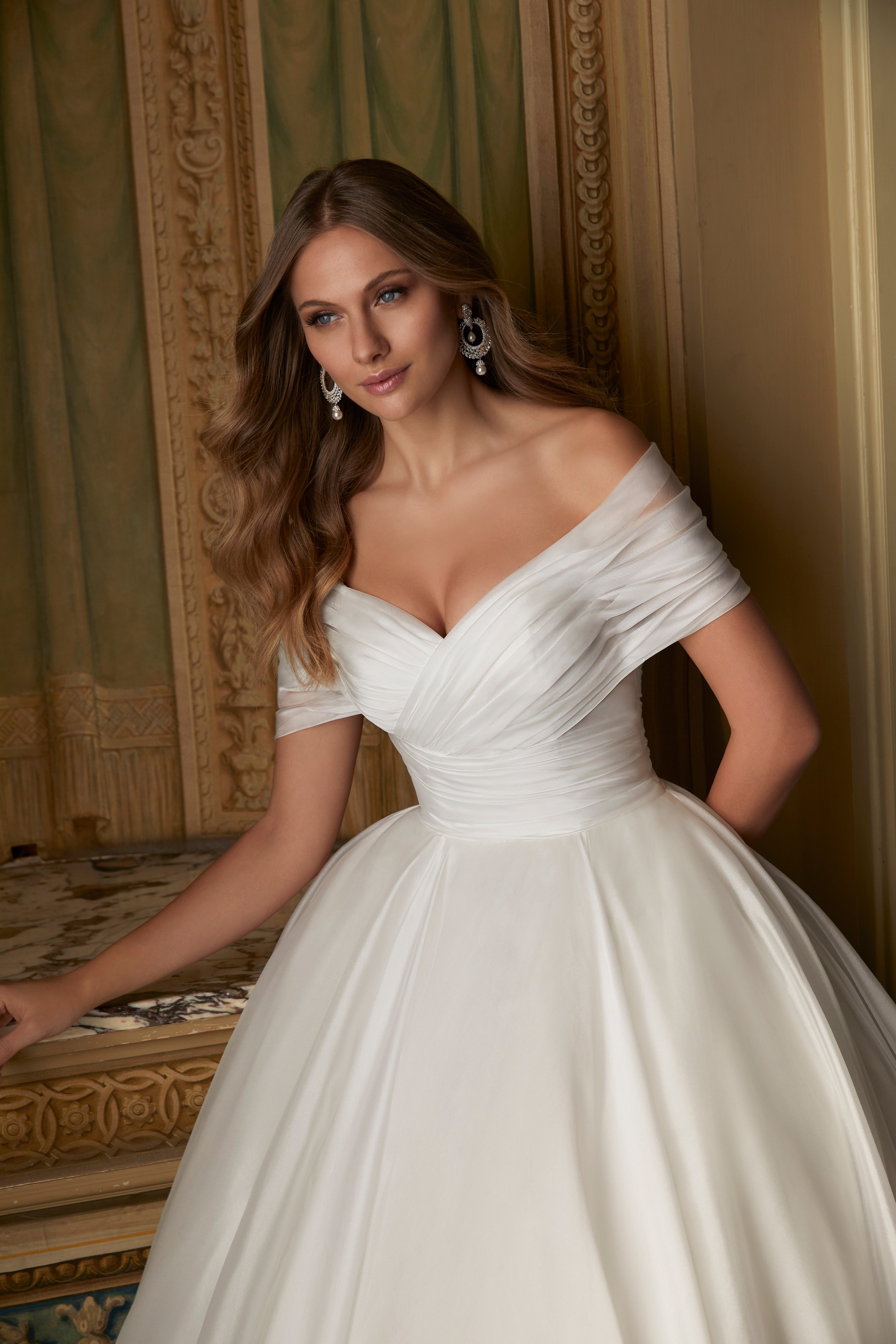 Close up of a model stood in a ballroom in a bardot style wedding dress (Ronald Joyce style 69551), a plain off-the-shoulder ballgown with a pleated bodice