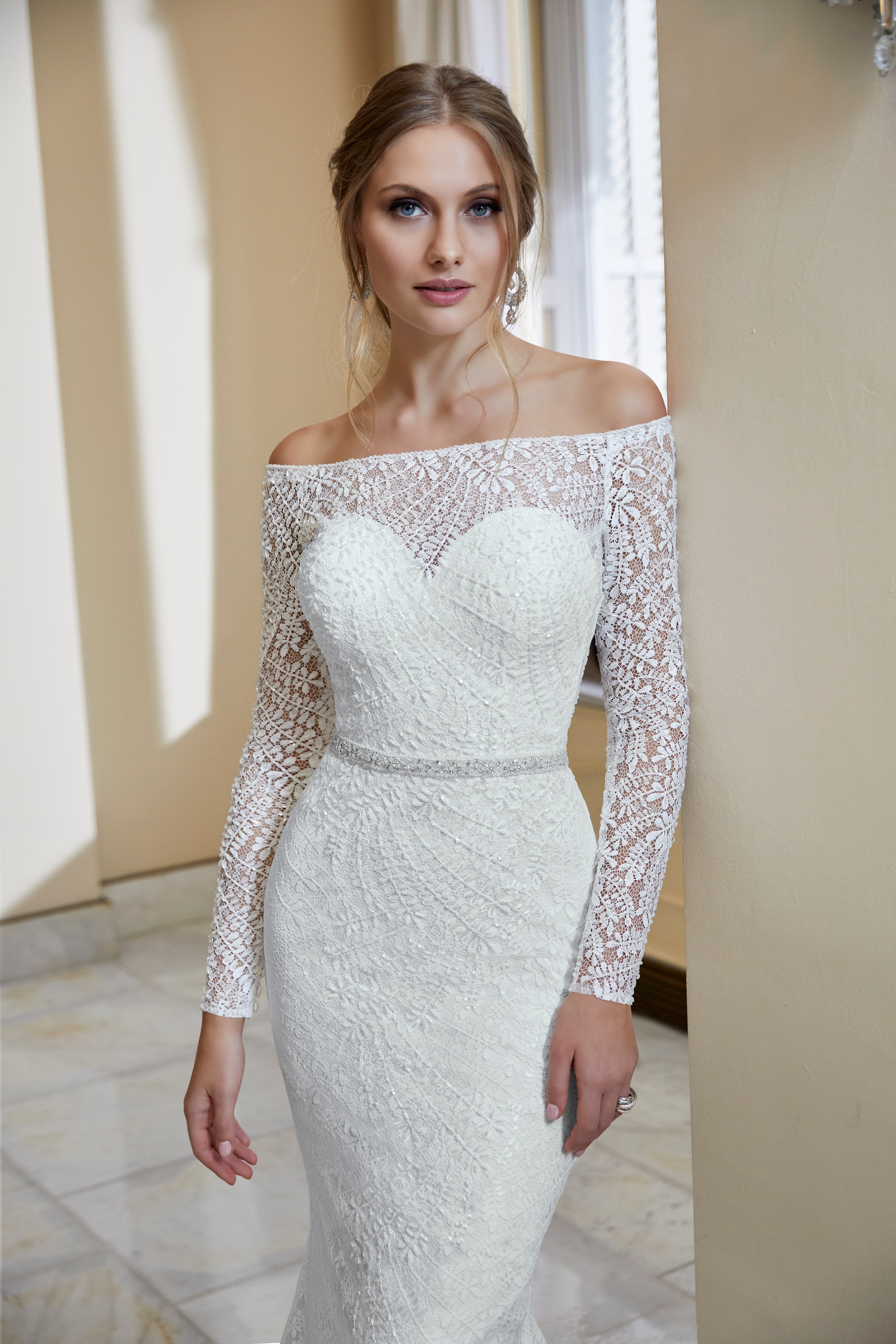 Model stood in a grand hallway in a bardot wedding dress (Ronald Joyce style 69568), a lace fit and flare wedding dress with a sheer sweetheart neckline and matching long sleeves
