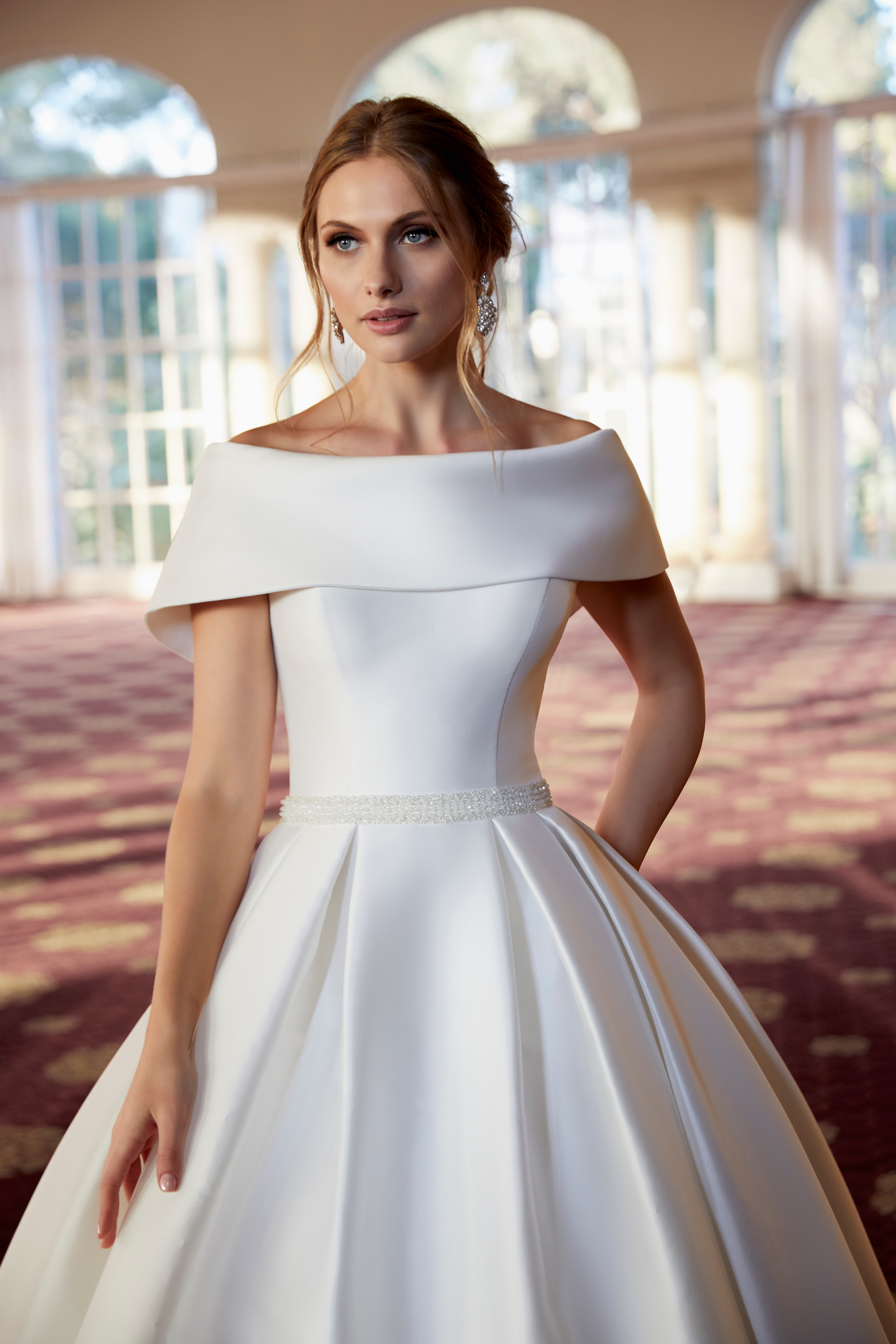 Model stood in a ballroom in a bardot wedding dress (Ronald Joyce 69570), a white ballgown with a striking off the shoulder sleeve and delicately beaded white belt