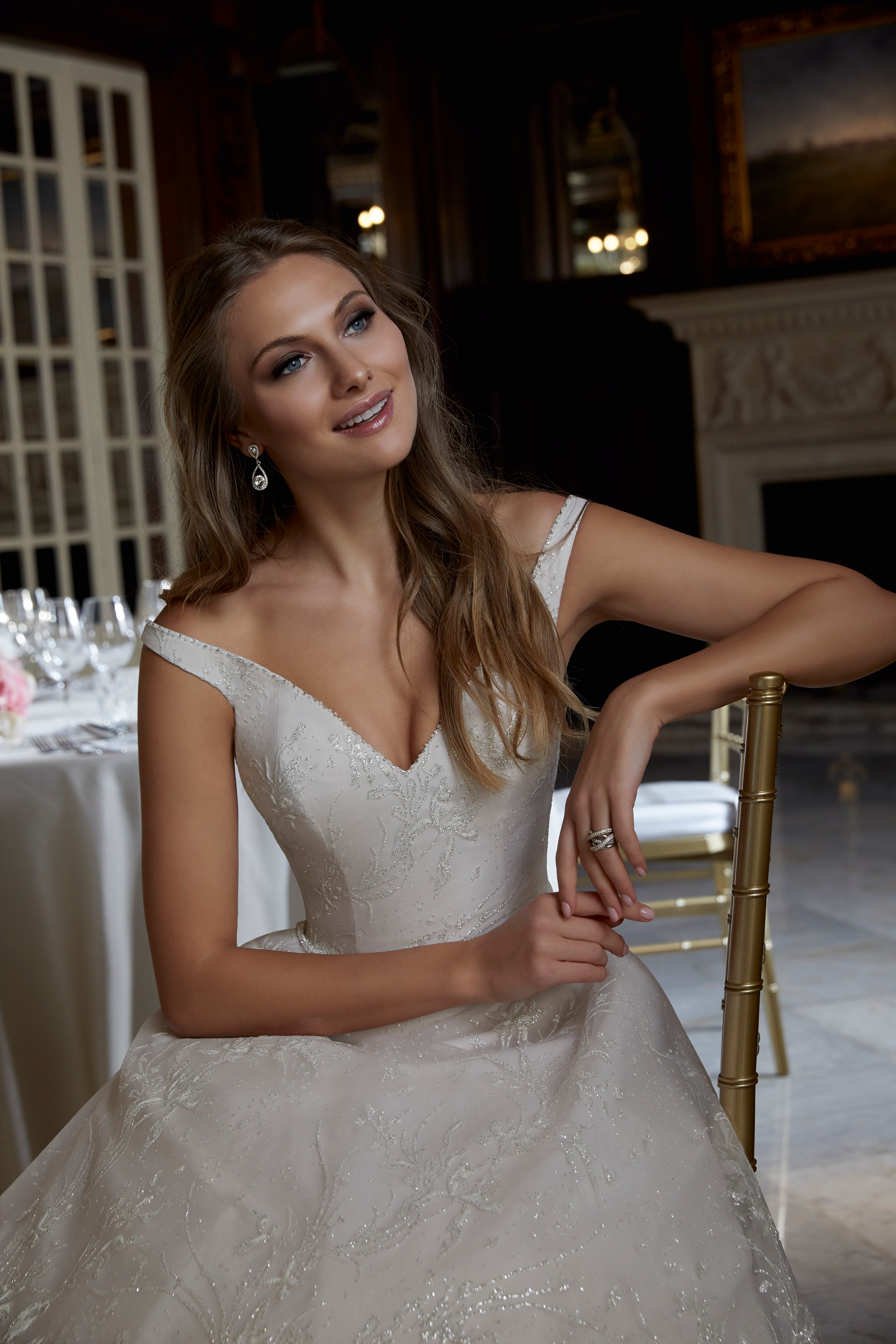 Close up of a model sat on a chair in a ballroom wearing a bardot wedding dress (Ronald Joyce 69582), an off-the-shoulder strappy ivory ballgown with silver beading and a v-neckline