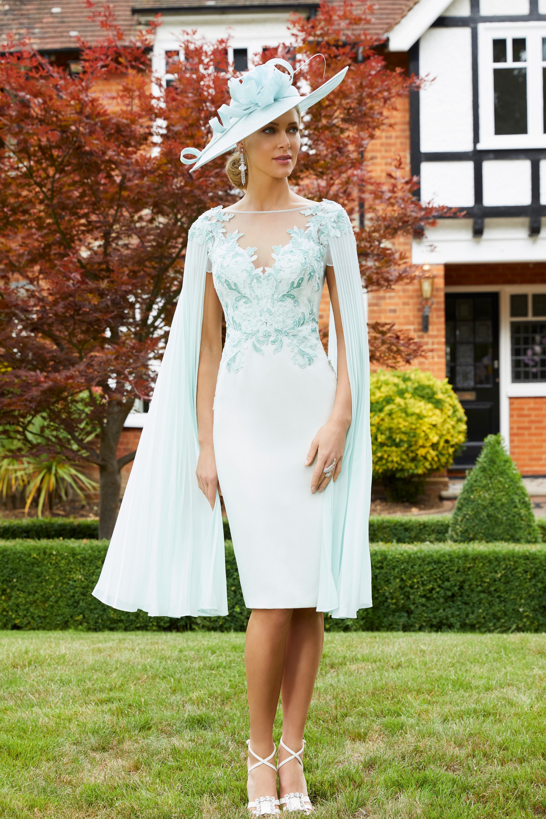 Model stood on a manicured lawn outside a house in Veni Infantino 991706, a elegant fitted knee-length dress with long pleated cape sleeves in aqua. 