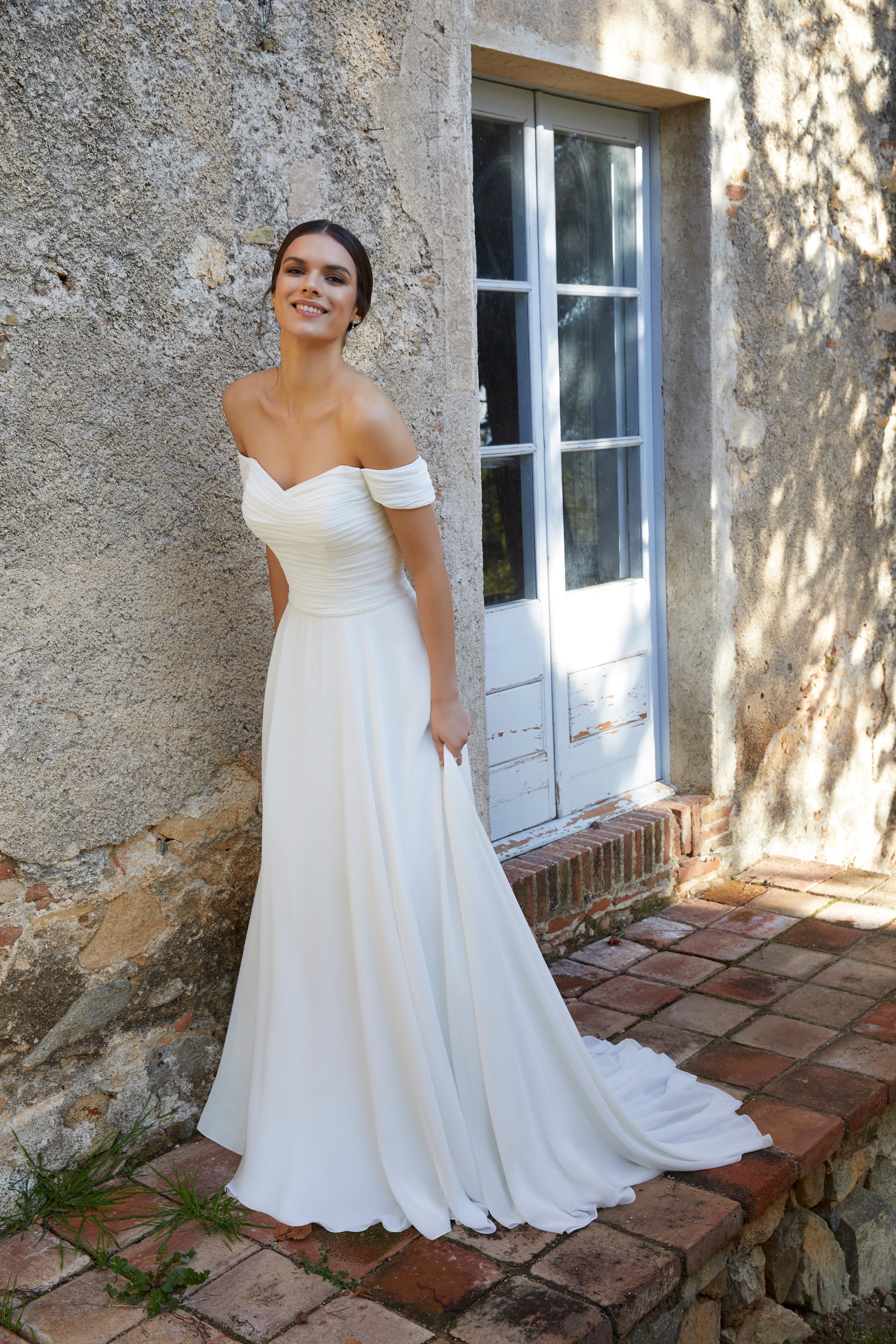 Model stood outside an old Italian building in Ronald Joyce style 18461, a chiffon sheath wedding gown with capped off-the-shoulder sleeves that’s perfect for small weddings