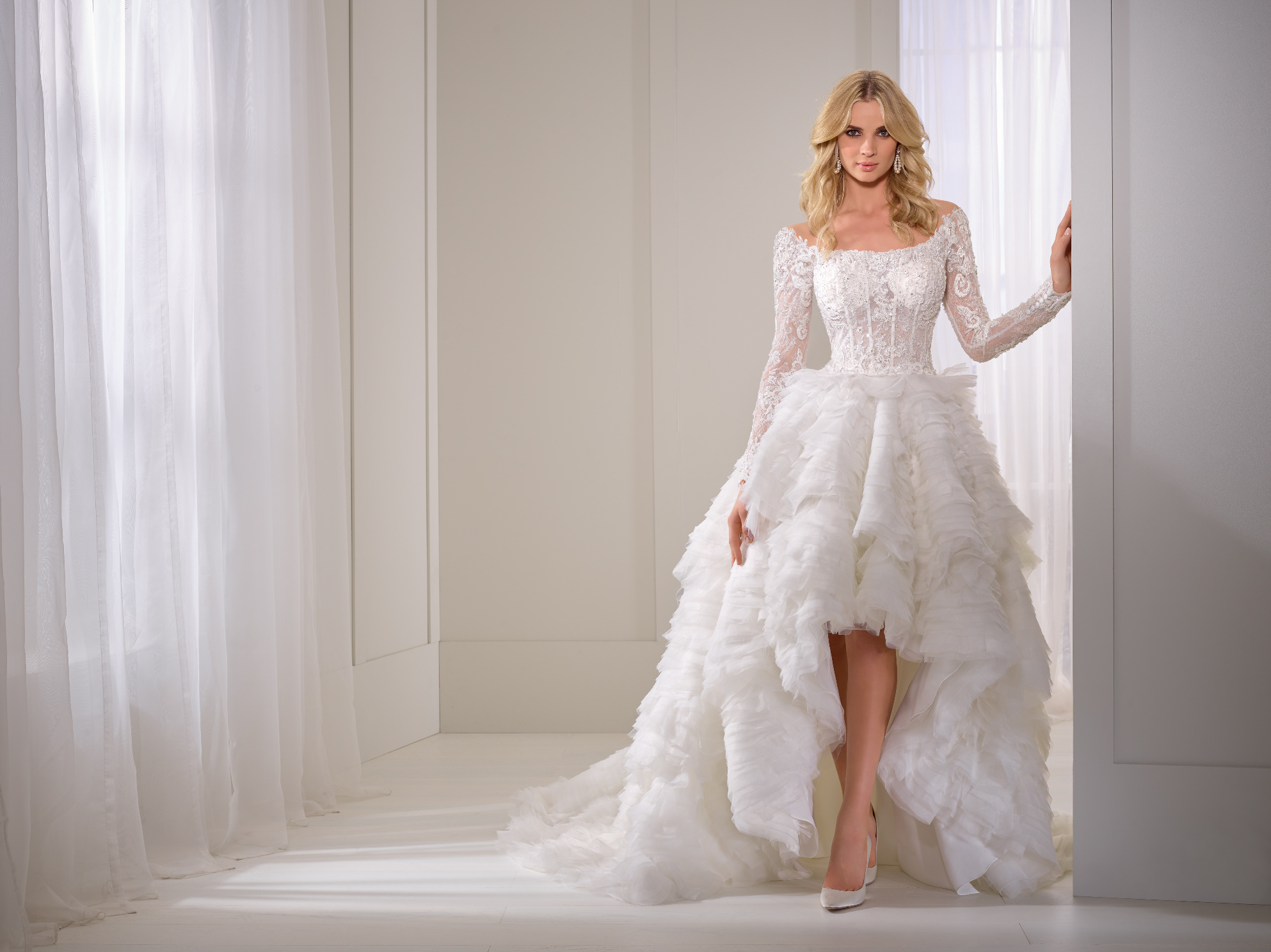 Photo of a model in Ronald Joyce 69375, a hi lo wedding dress silhouette with a lace illusion bodice and matching long sleeves complete with an organza and tulle ruffled skirt