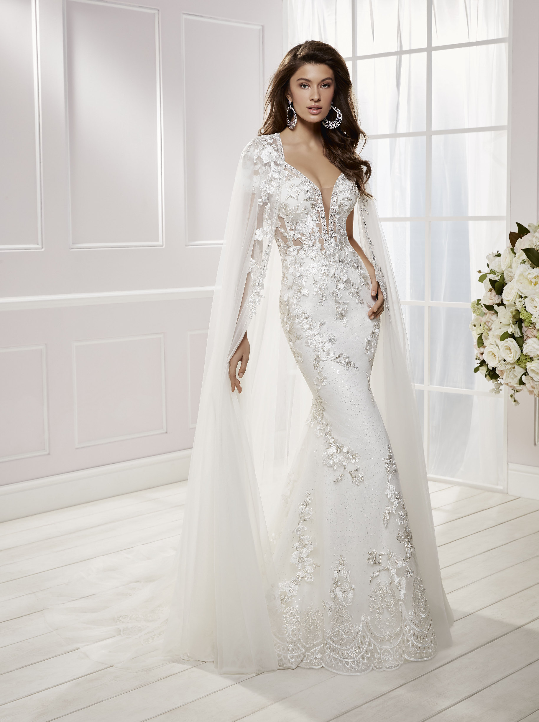 Model stood in a white room near cream flowers in Ronald Joyce 69454, a floral applique fit and flare wedding dress with cap sleeves, a plunging neckline, sparkle trim and cape