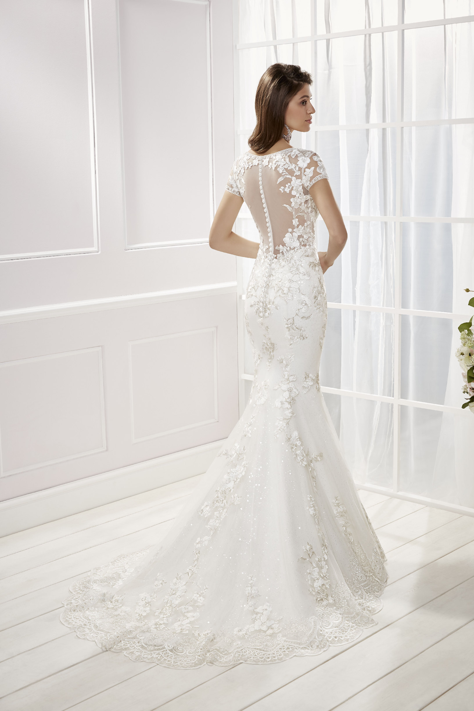 Back of a brunette model stood in a white room in Ronald Joyce 69454, a floral applique fit and flare wedding dress with a bridal buttoned illusion back, cap sleeves and a lace hem