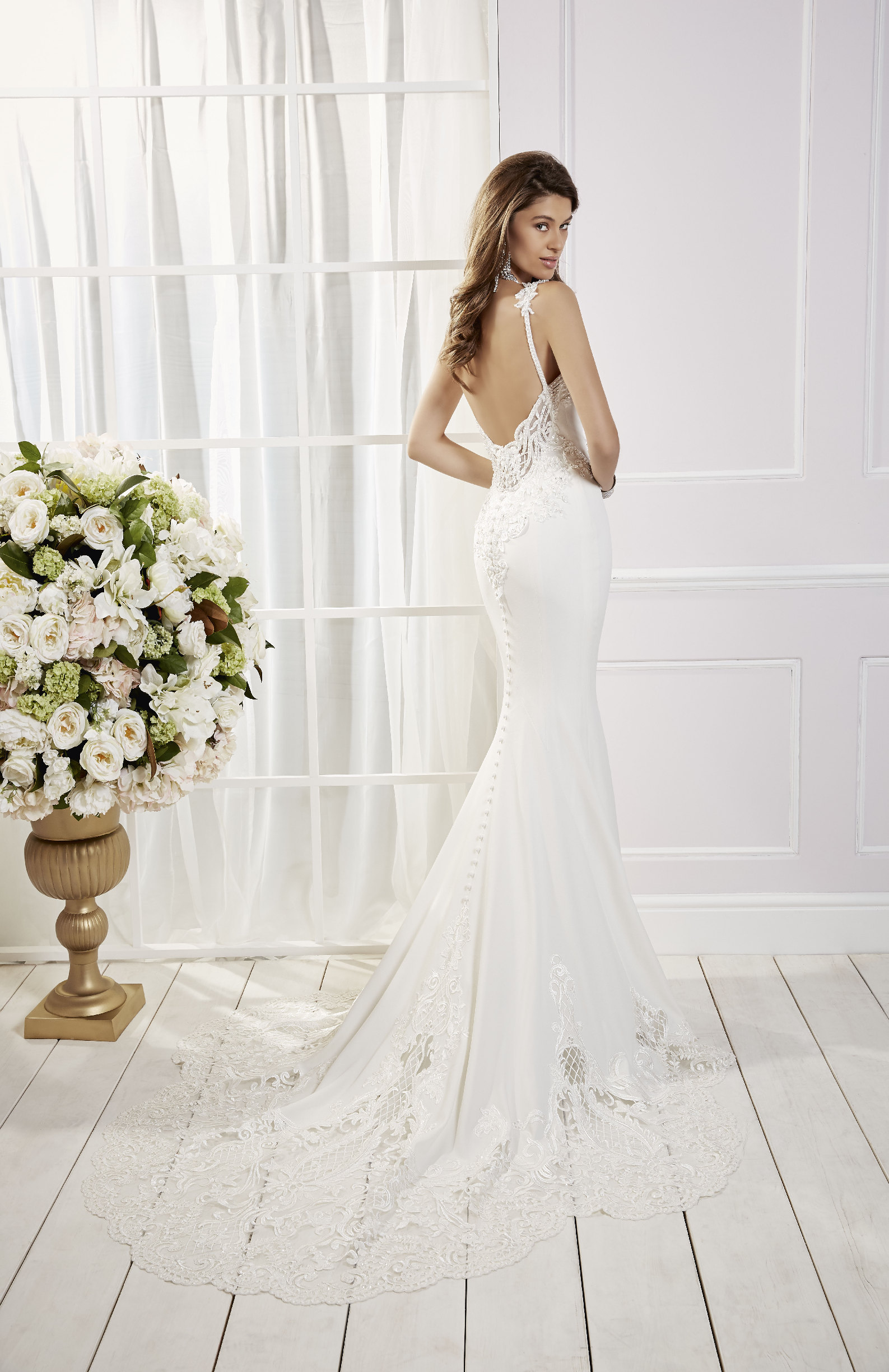 Model stood in a white room by cream flowers in Ronald Joyce 69458, a fishtail wedding dress with delicate straps, an open back, bridal buttons, a plain skirt and lace scallop hem